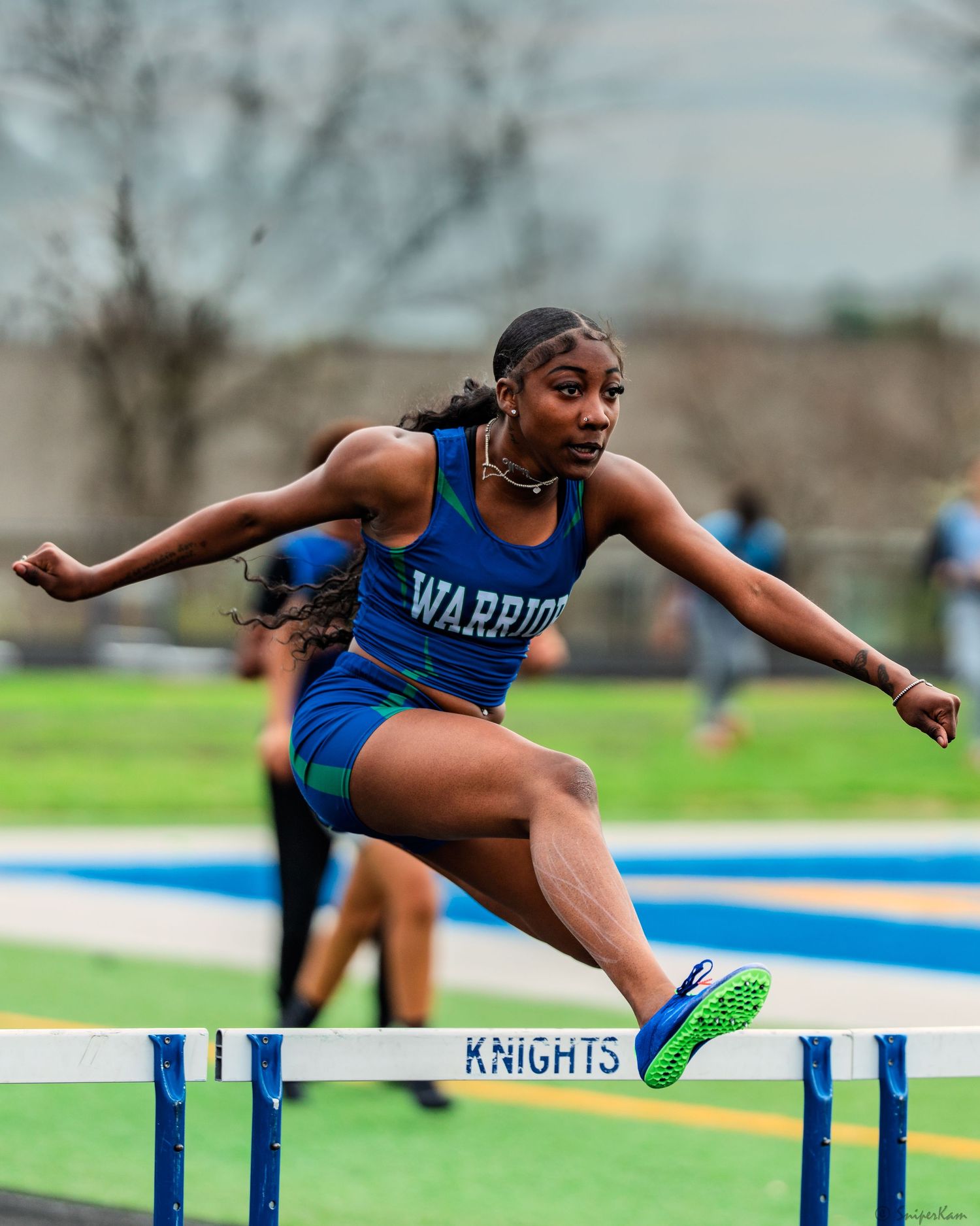 Female Warriors track athlete clears hurdle during competitive high school track meet.