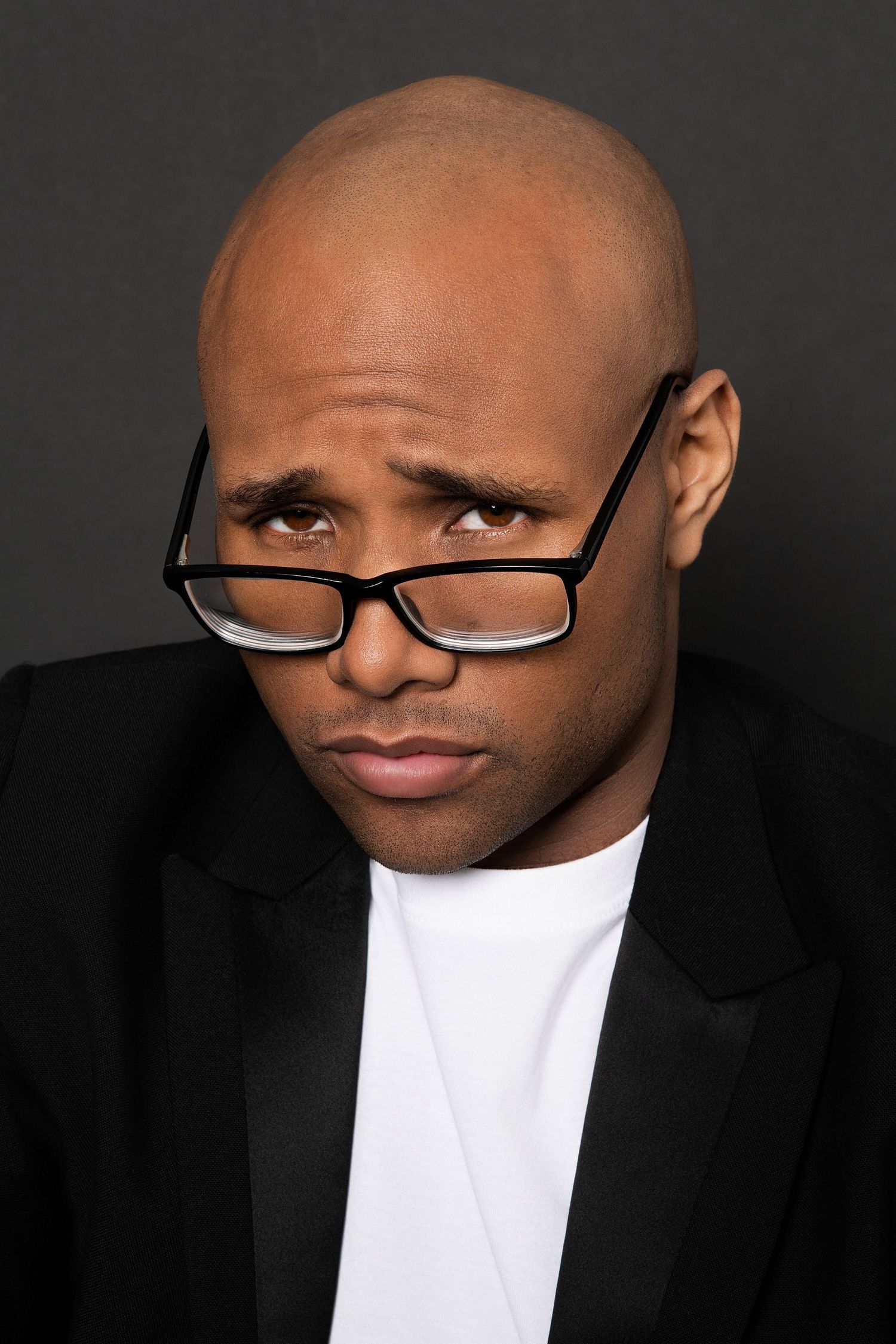 Bald Black man in black blazer and white shirt looking intensely over rectangular glasses against dark background.