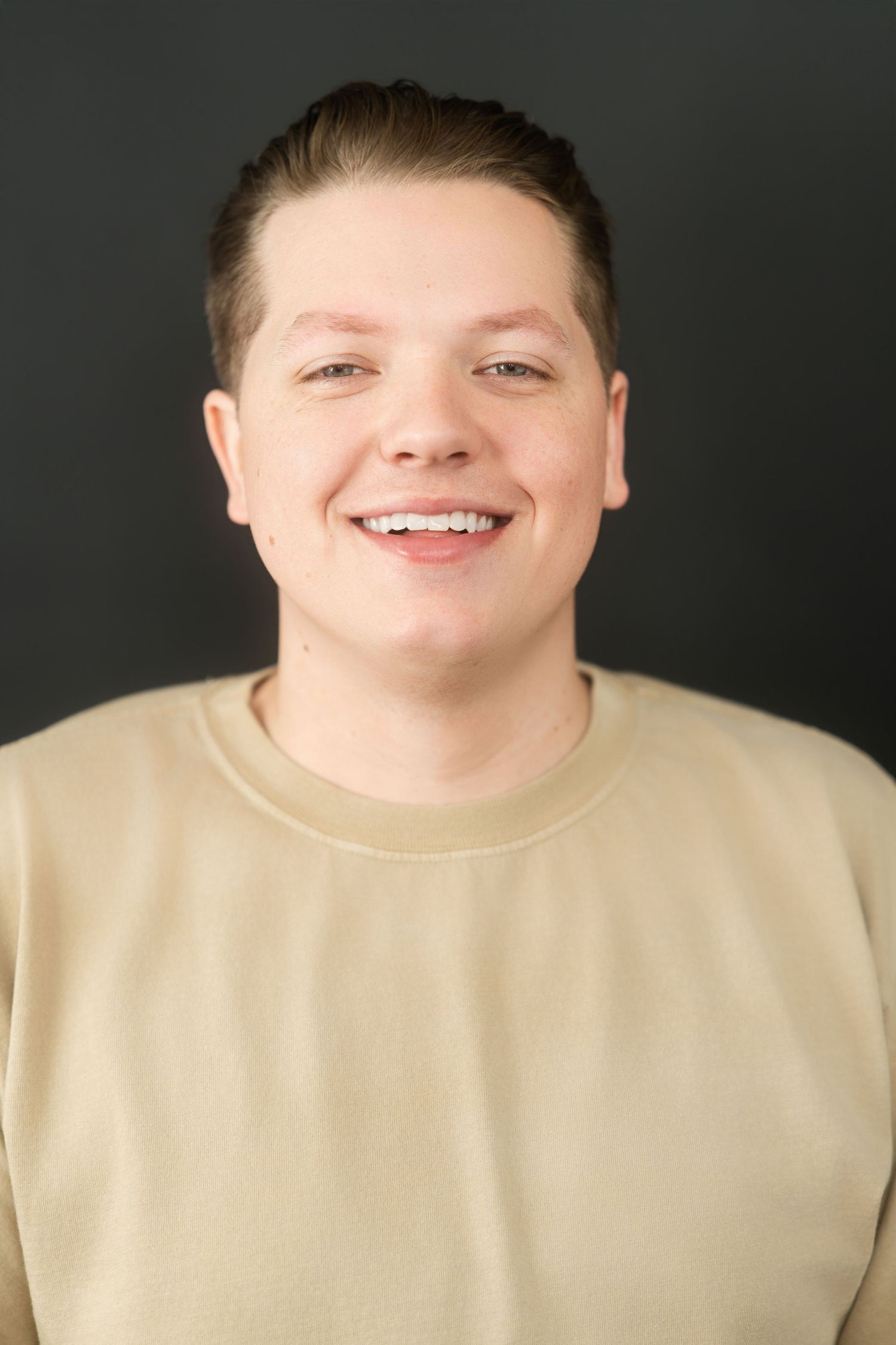 Young man in beige sweatshirt smiling broadly in studio portrait against dark background.