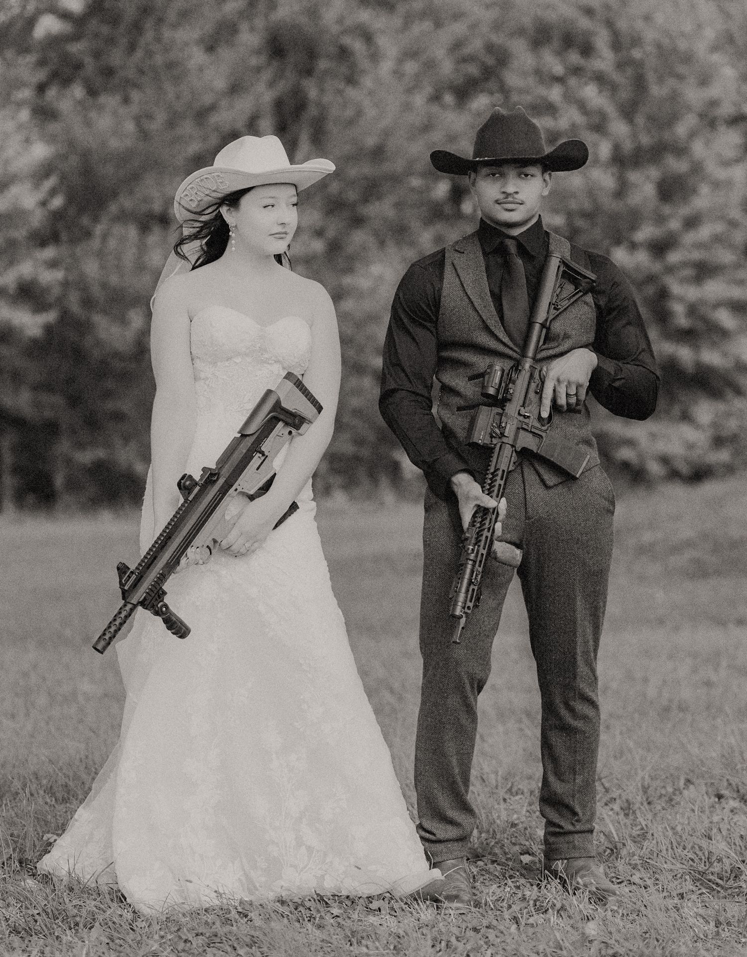 Black and white wedding photo of bride and groom in cowboy hats holding rifles.