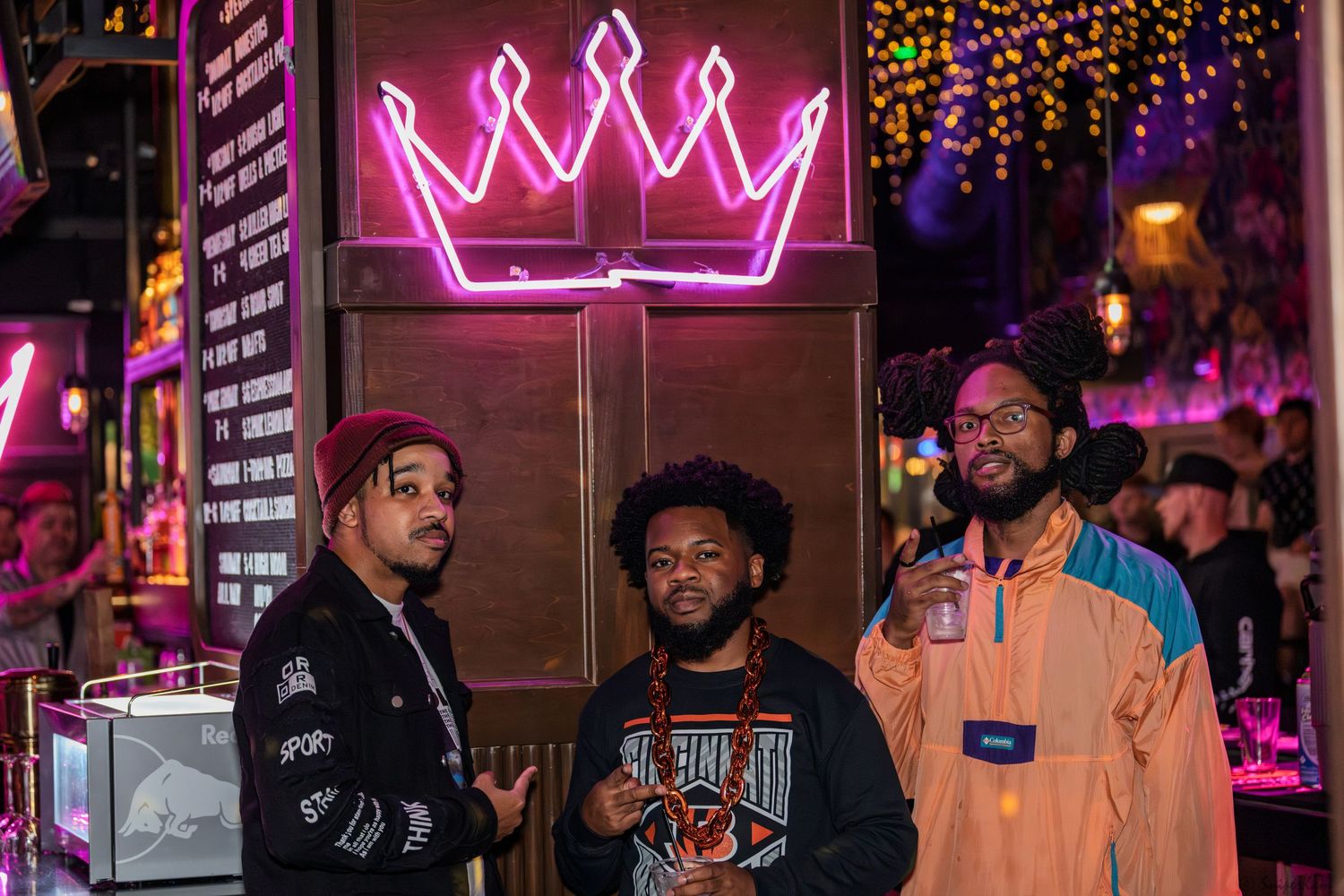 Three men posing together under neon crown sign at vibrant nightlife venue.