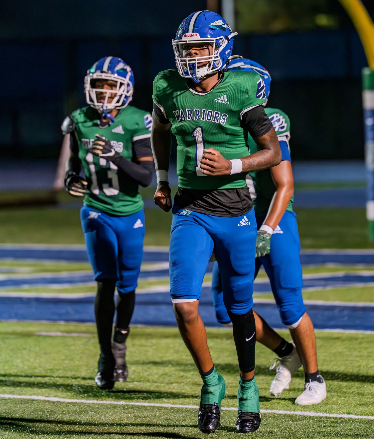 High school football player number 1 in green and blue Warriors uniform jogging on field during game.