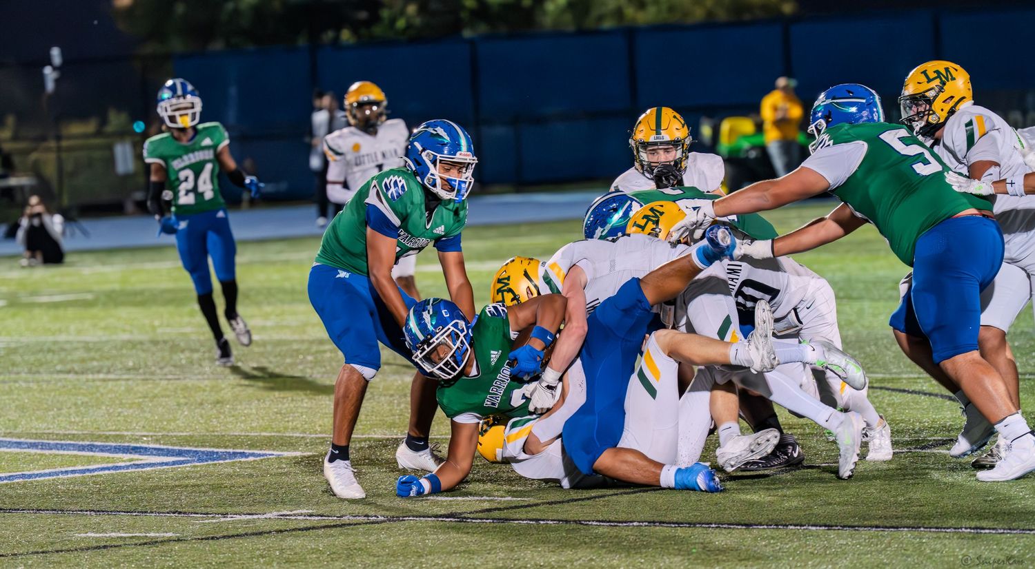 Warriors players in green and blue uniforms tackle opposing players in yellow during a football game.