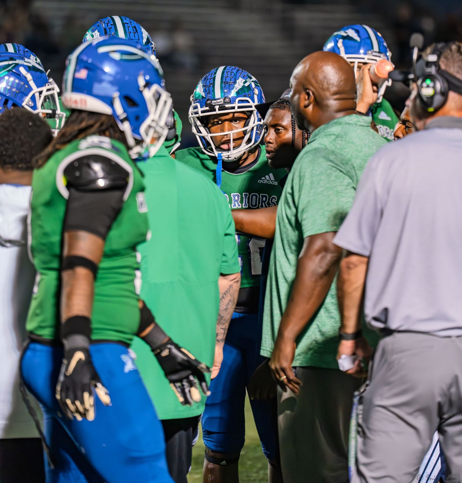 Football players in blue and green uniforms huddle with coaches on the sideline at night.