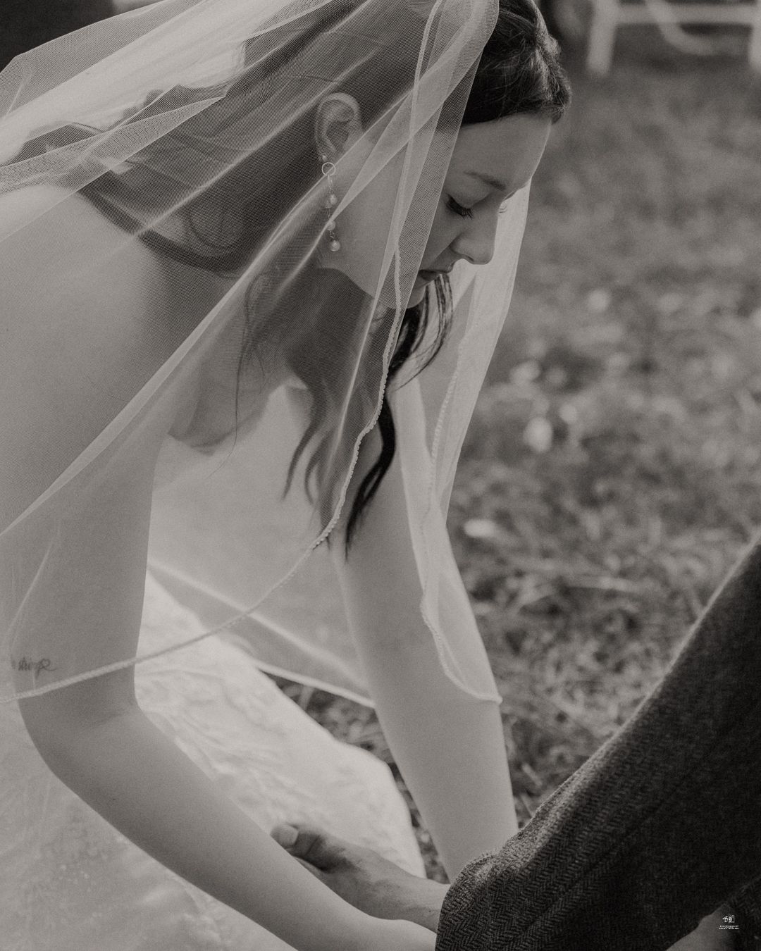 Black and white artistic shot of veiled bride looking down while holding grooms hand outdoors.
