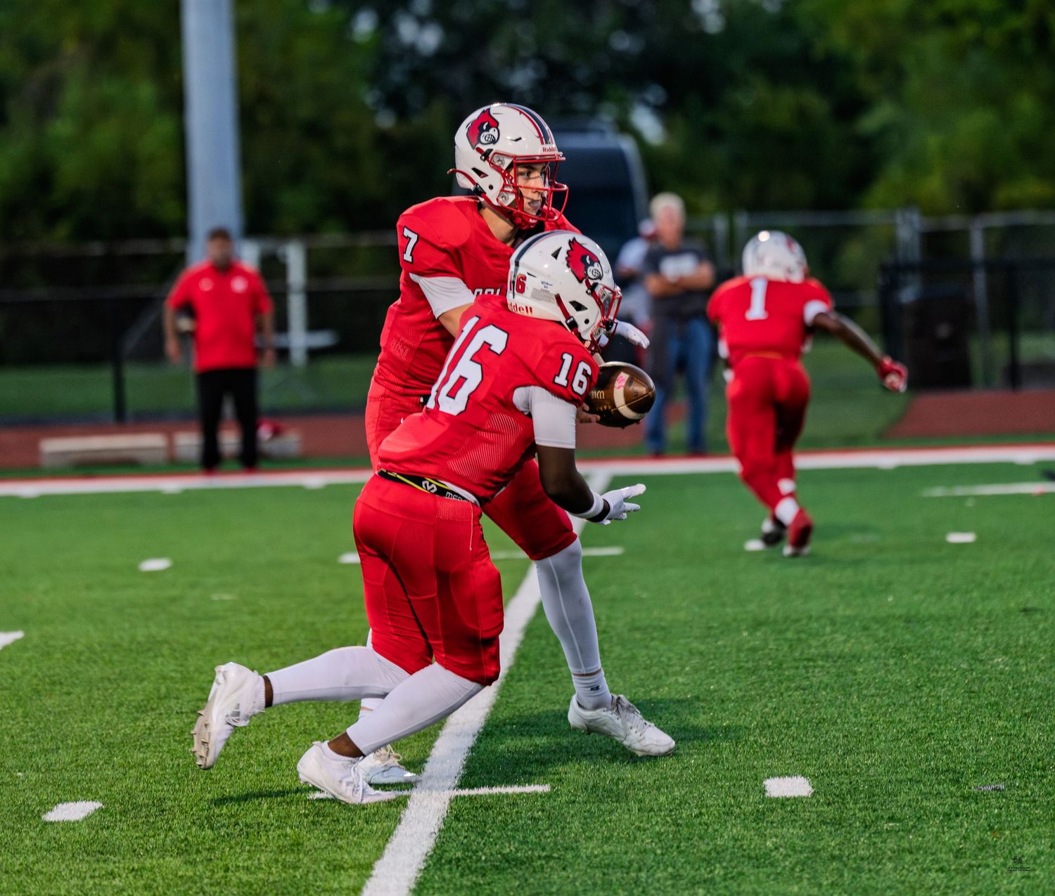 Red team quarterback #7 hands ball to #16 during a high school football game at dusk.