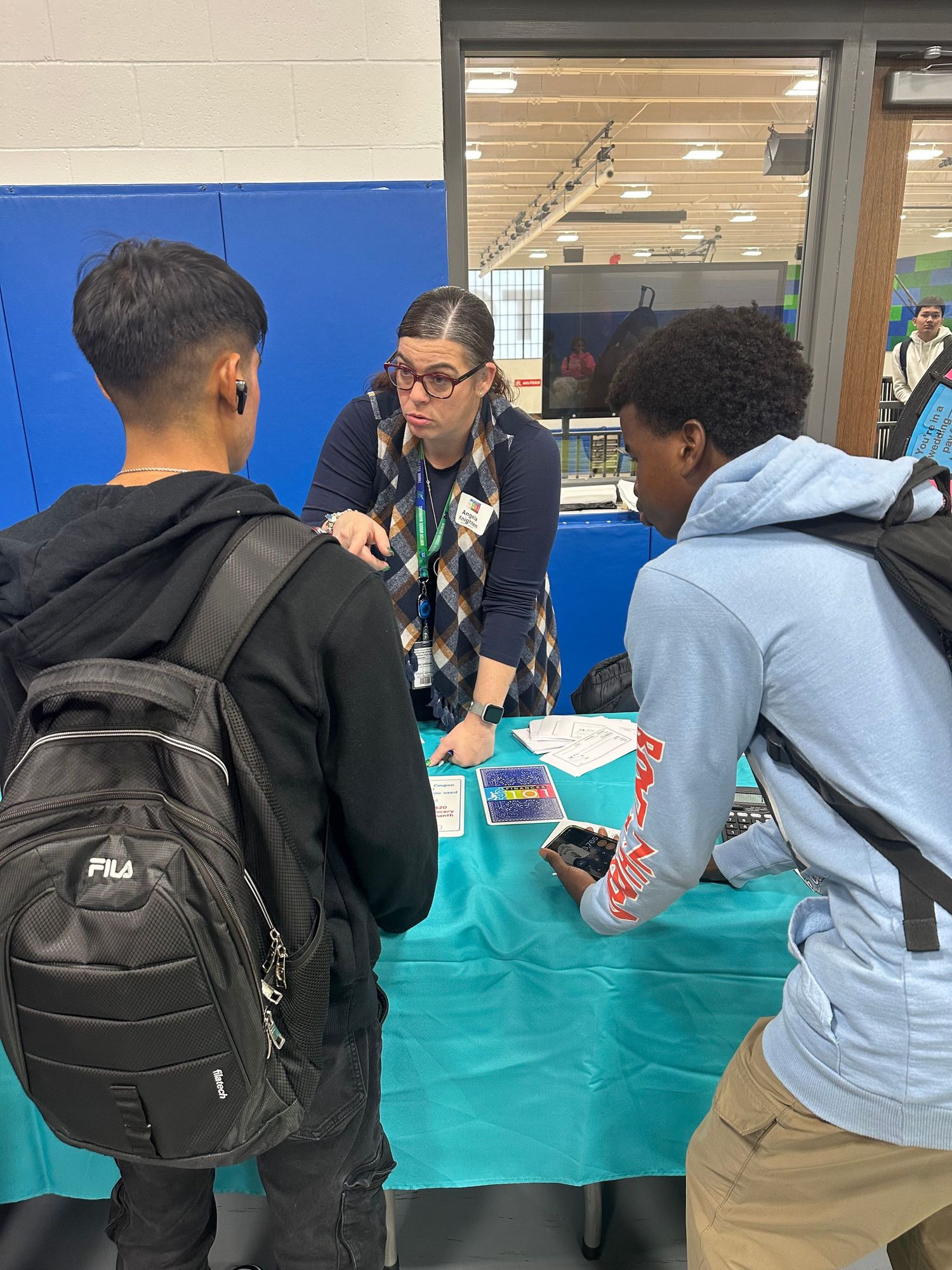 A teacher with glasses assists two students at a teal table during a school fair event in a gymnasium.
