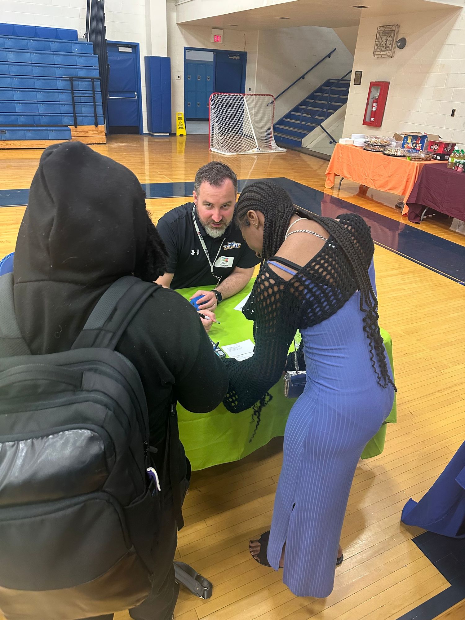 Adults and students gather around a table in a school gymnasium during an educational event or fair.