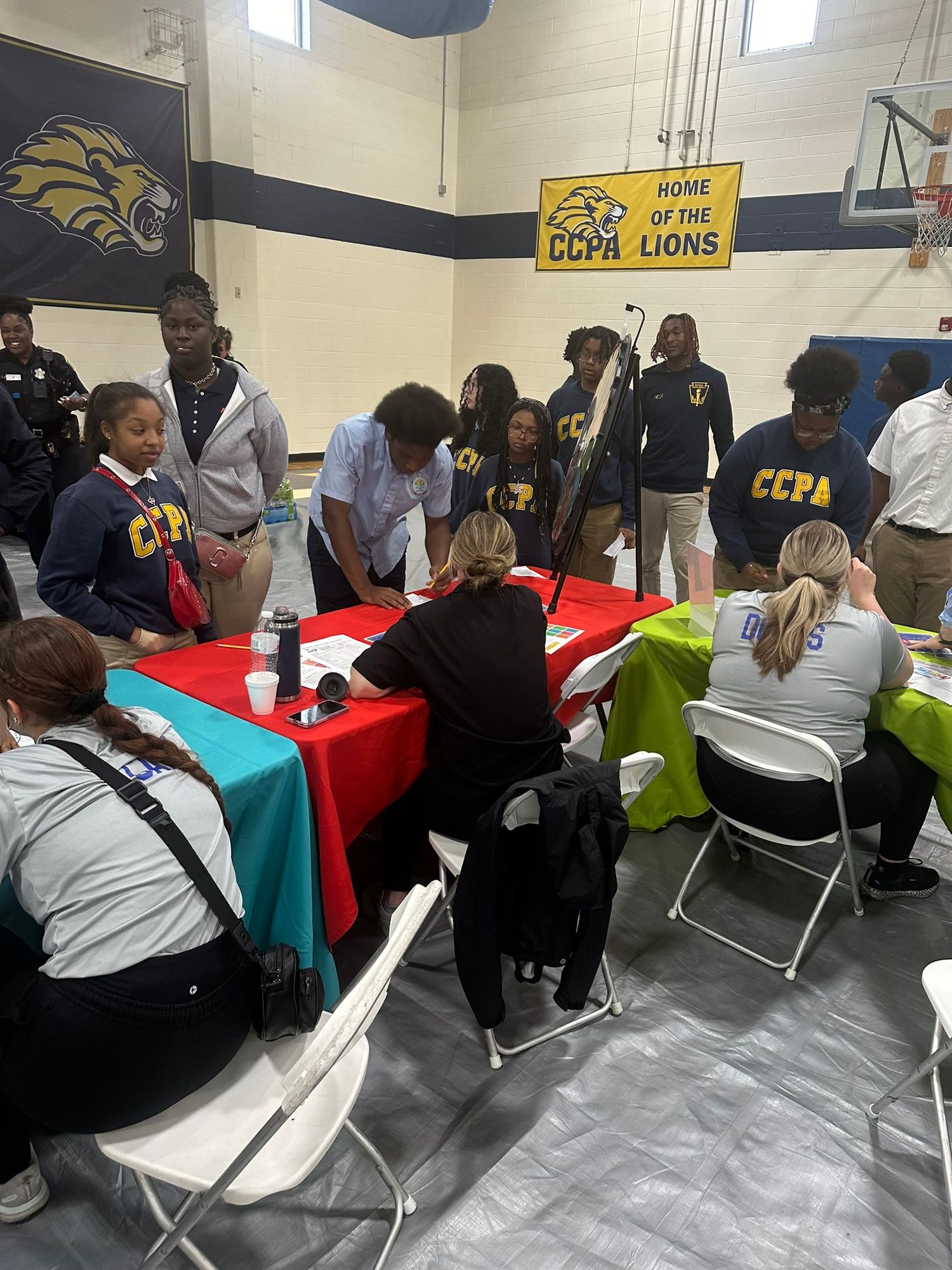 Students in CCPA uniforms gather around a table in a school gymnasium decorated with a Lions banner.