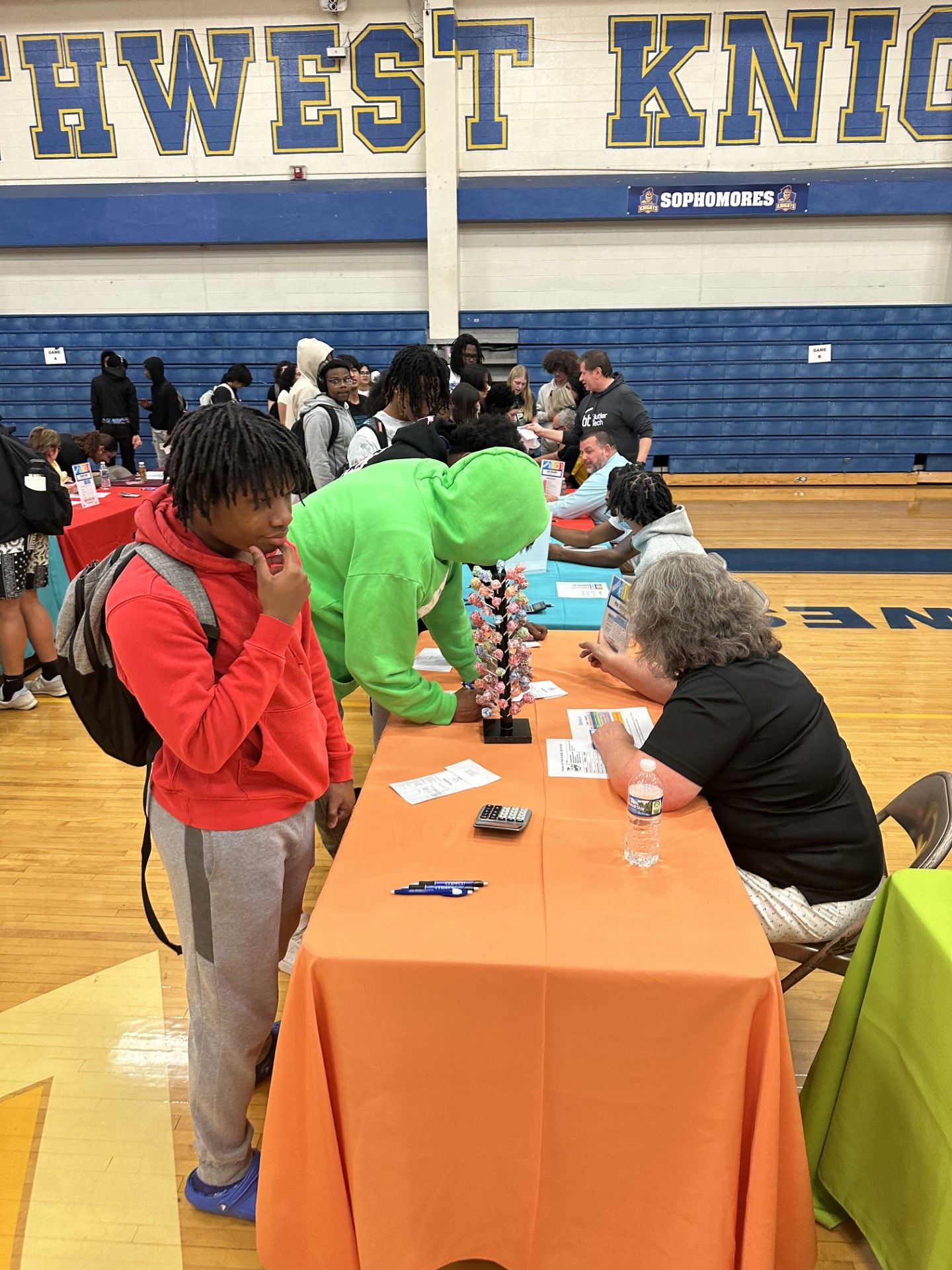 Students gather around an orange-draped table at a Southwest Knights high school gymnasium career or college fair event.