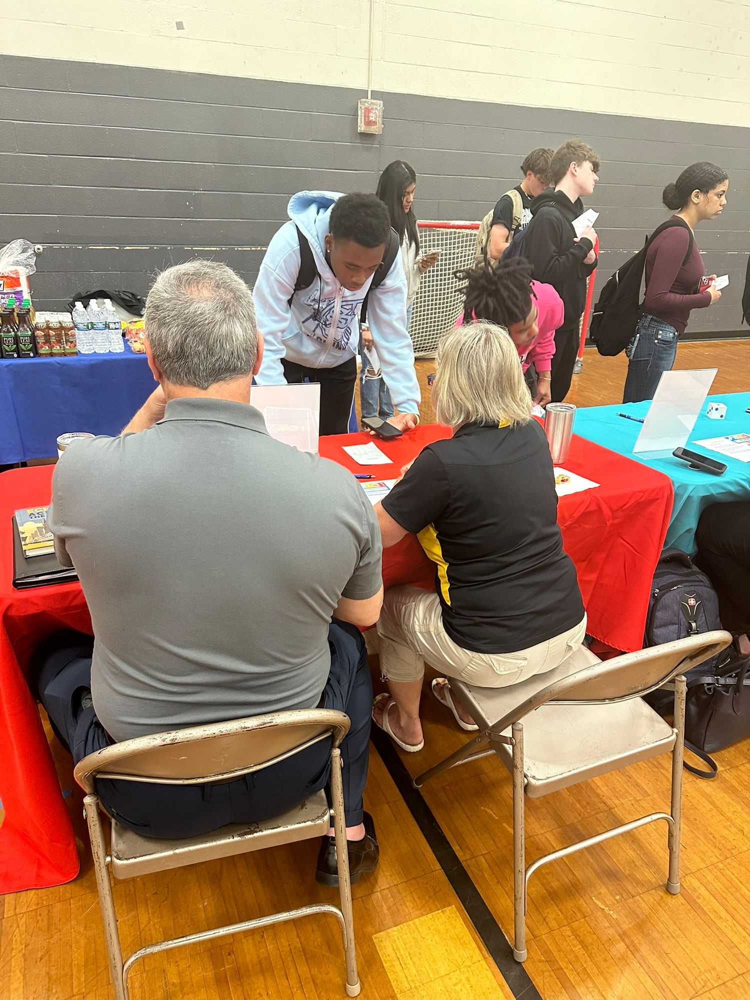 Students and adults interact at a school career fair with colorful tables in a gymnasium.