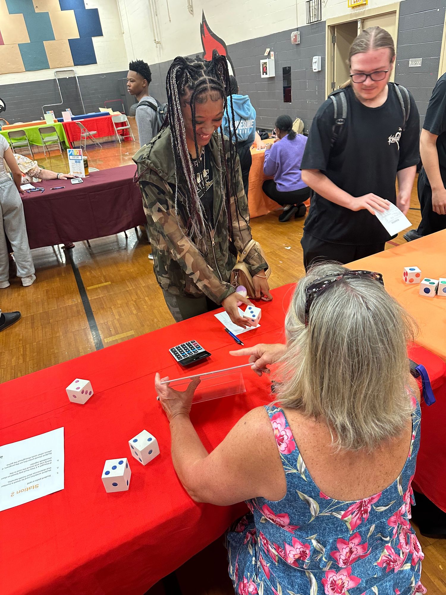 Students and teacher interact at a red table with dice during an educational fair in a school gymnasium.