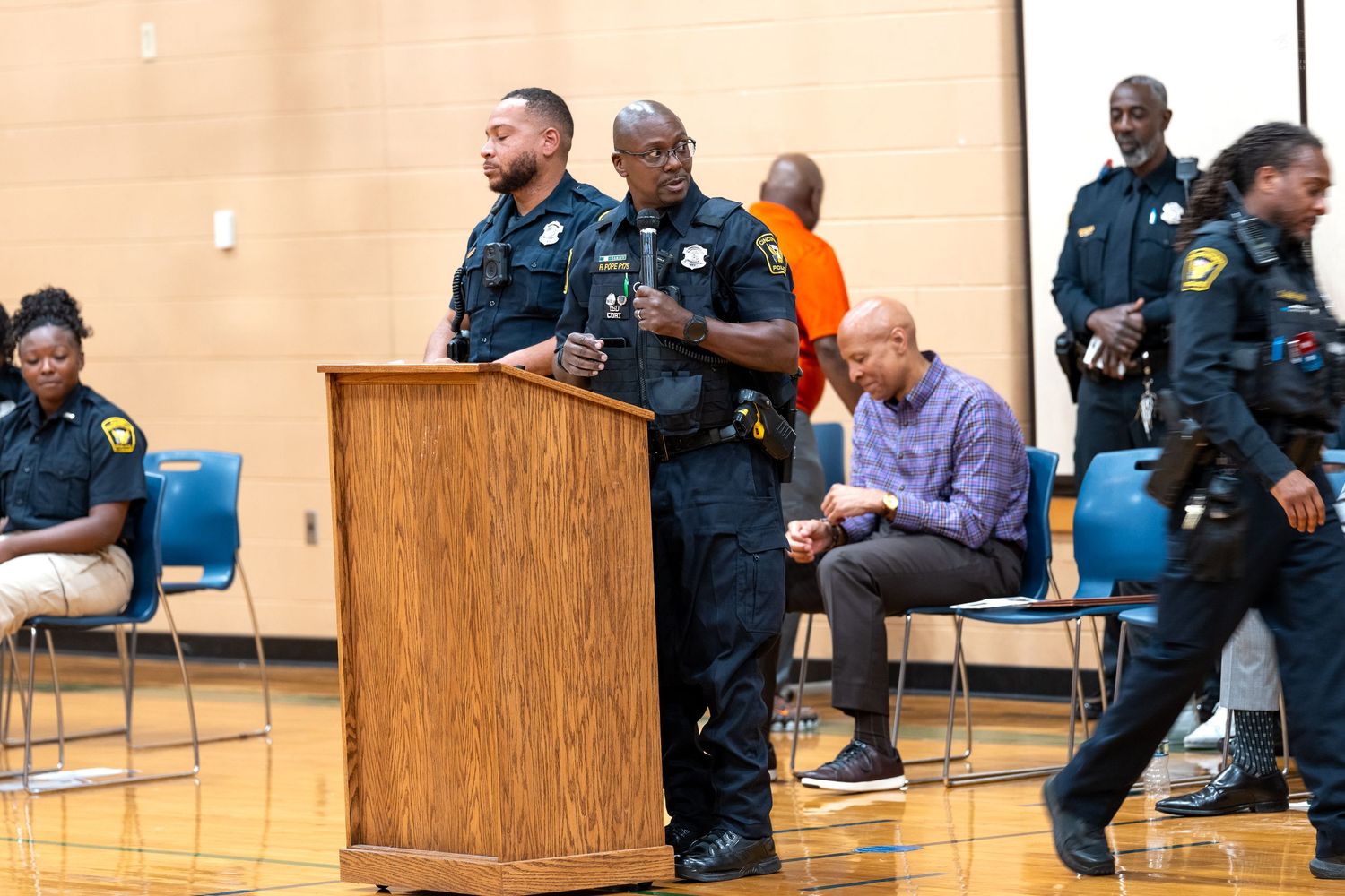 Two police officers stand at a wooden podium addressing a community meeting in a school gymnasium.