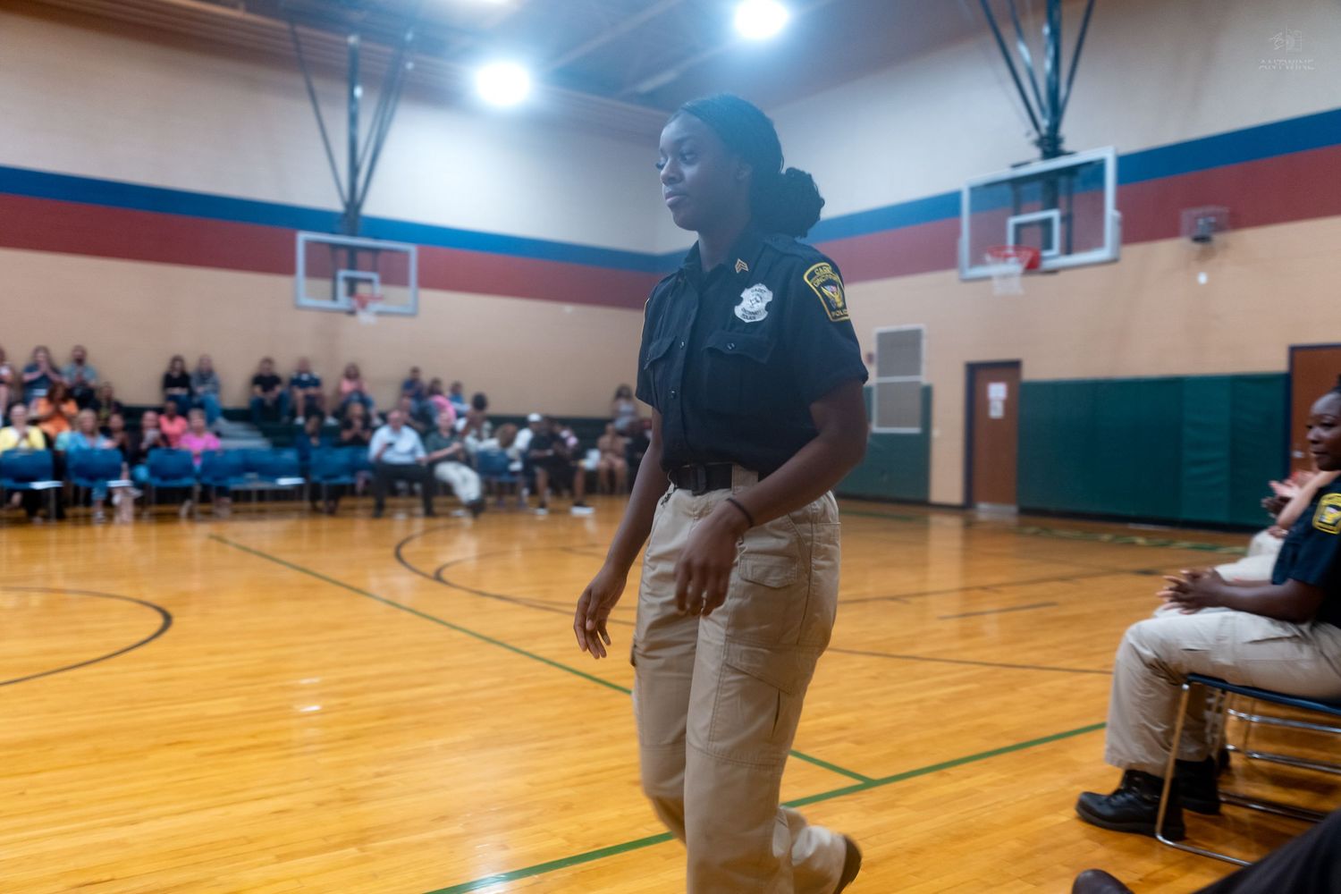 Female law enforcement officer in uniform stands in a school gymnasium during a community meeting.