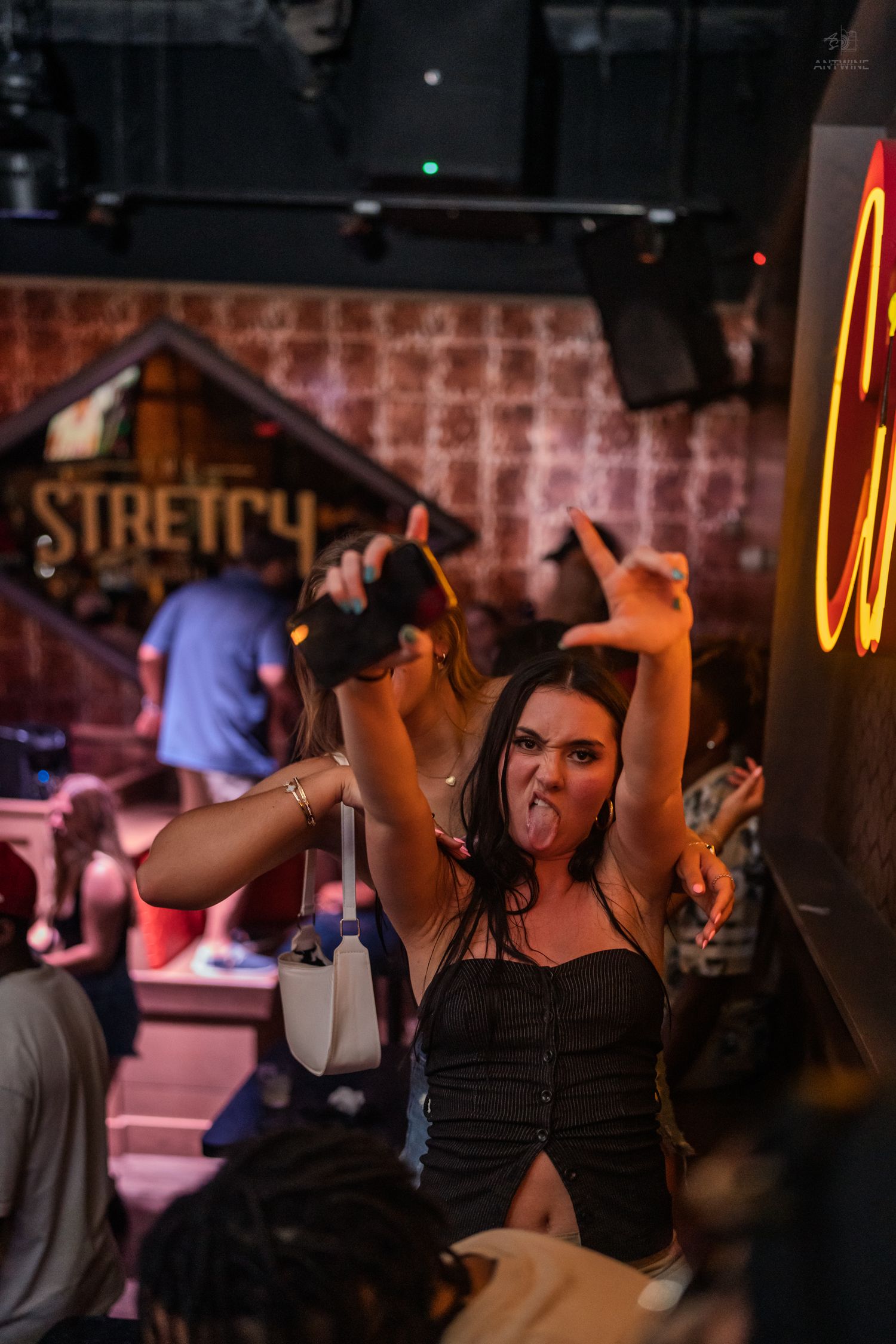 Excited woman sticking out tongue while holding drink near neon sign at nightclub.