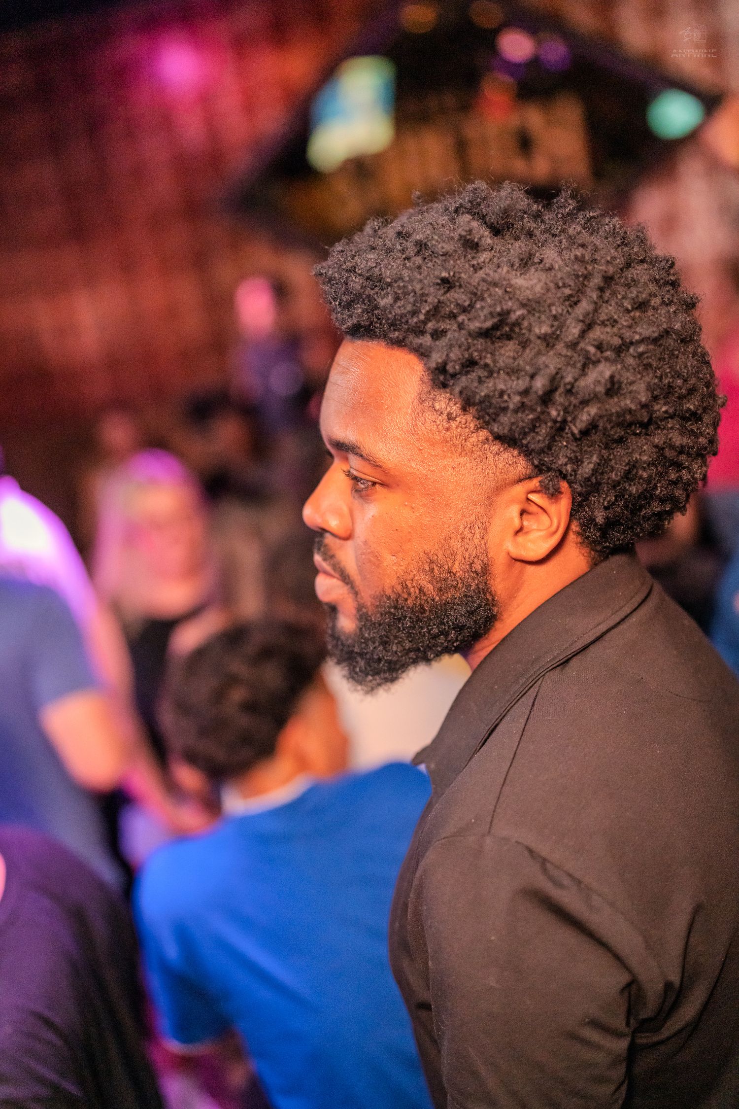 A Black man with a natural afro and beard in side profile at a lively nightclub setting.