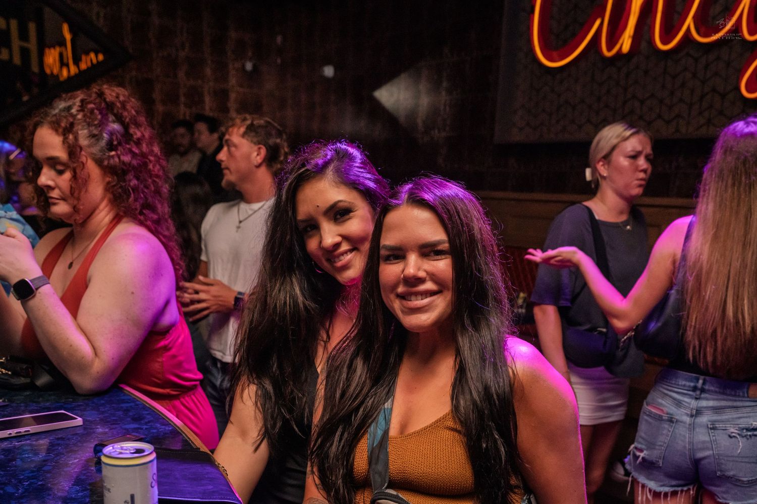 Two women smiling together at a lively bar with neon signs in background.