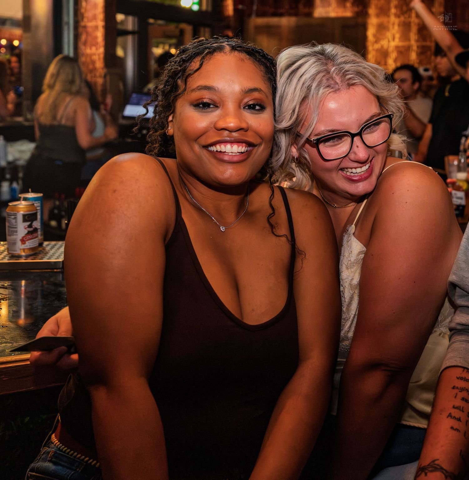 Two smiling women pose closely together at a lively bar with drinks visible in background.