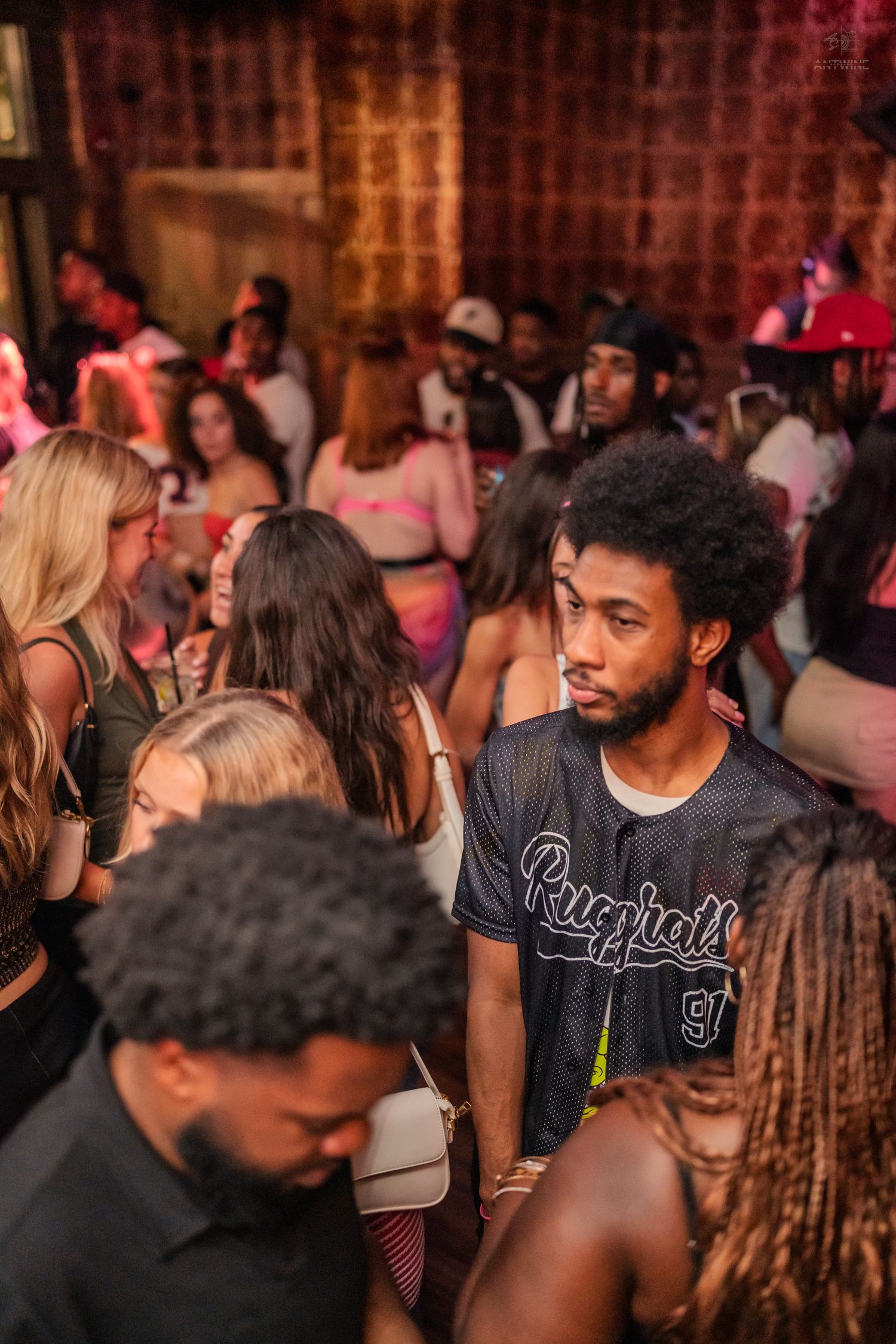 Young man in a Rugrats jersey standing among a diverse crowd at a nightclub.