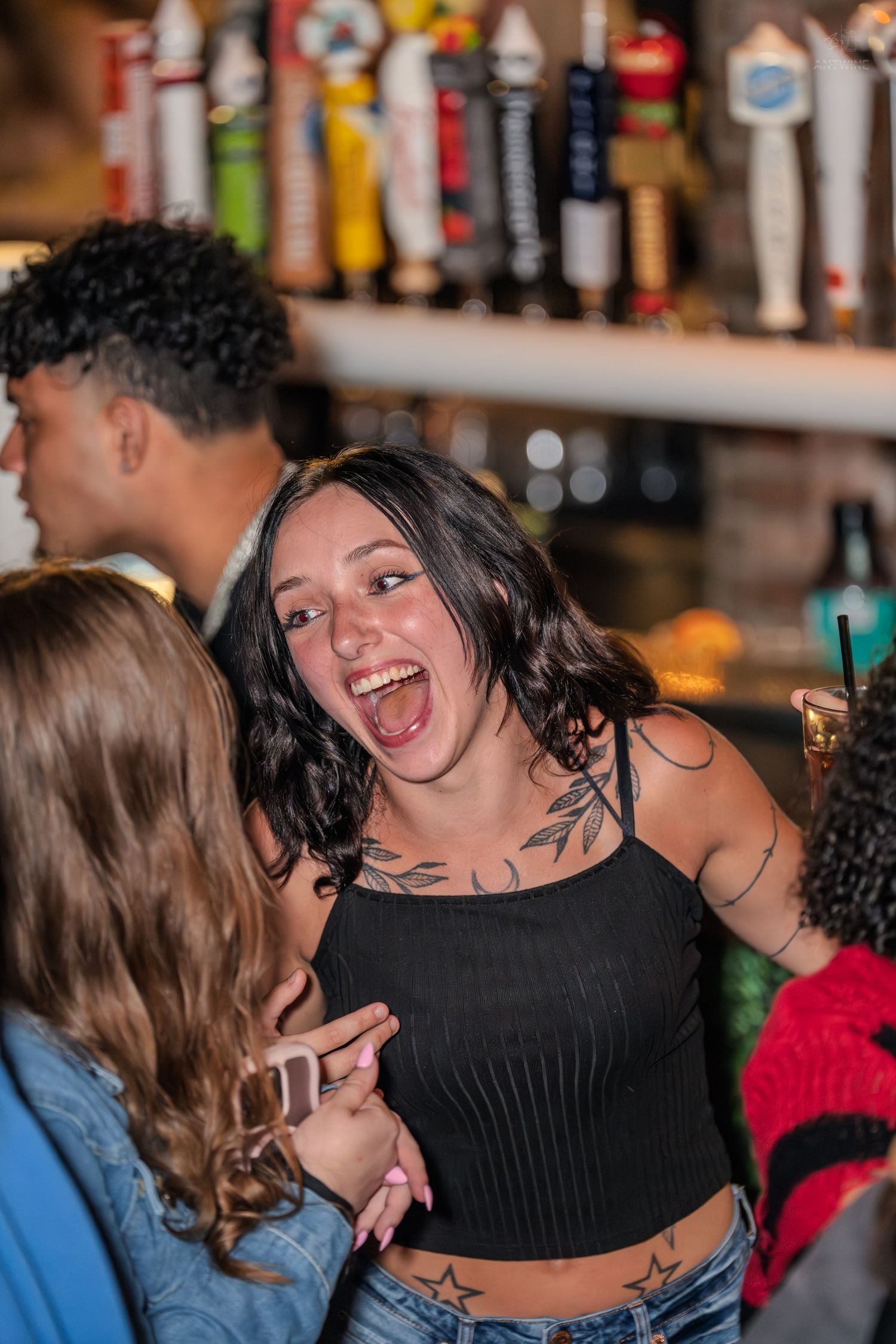 Laughing tattooed woman in black crop top socializing with friends at a lively bar.