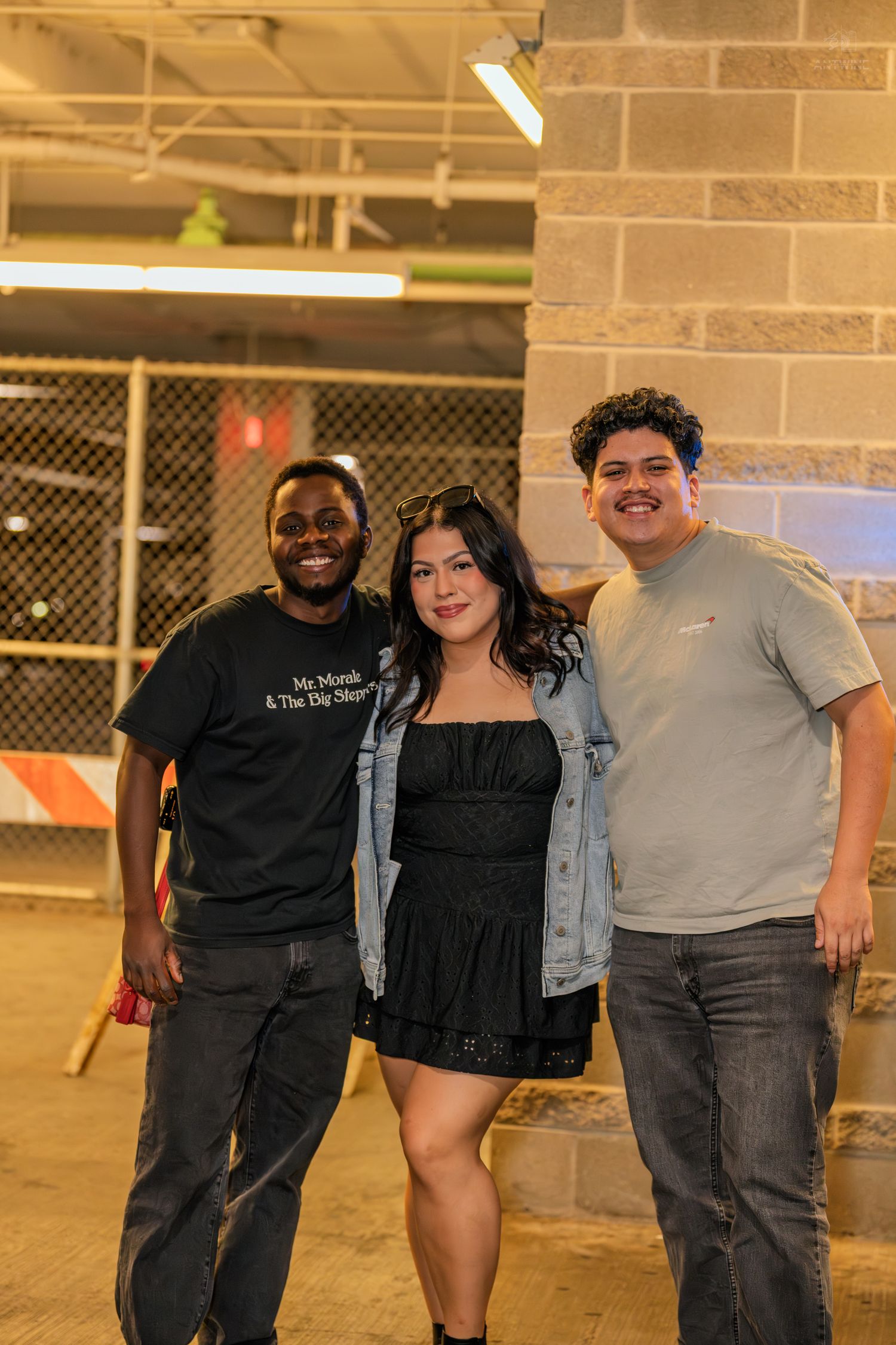 Three smiling friends posing together in a warmly lit parking garage during a nighttime outing.