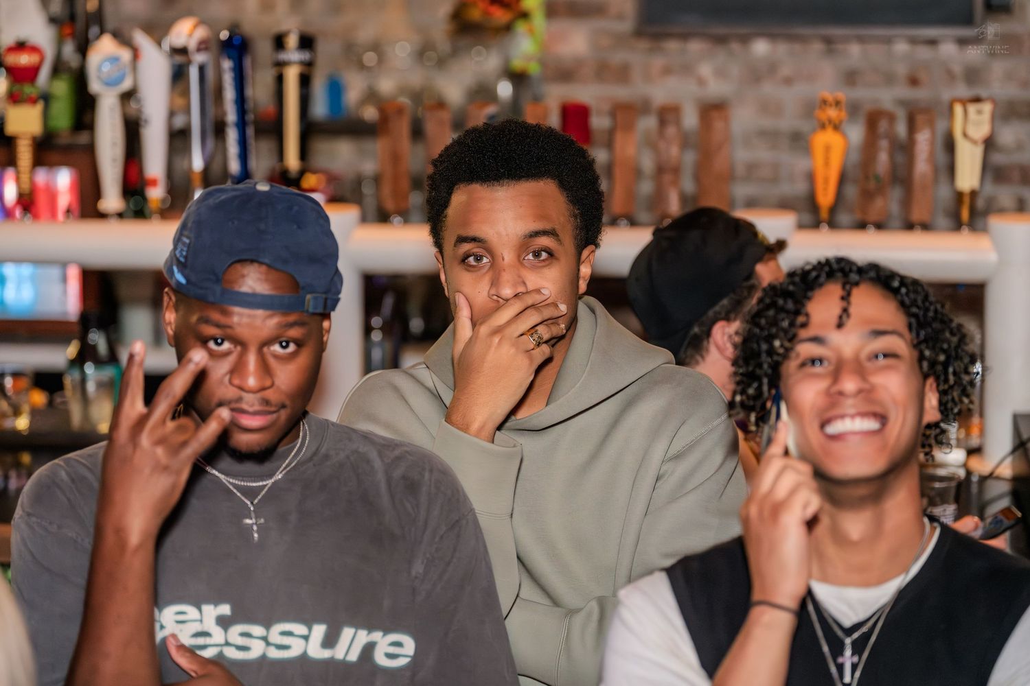 Three young men posing playfully at a bar, smiling and making hand gestures with tap handles behind them.