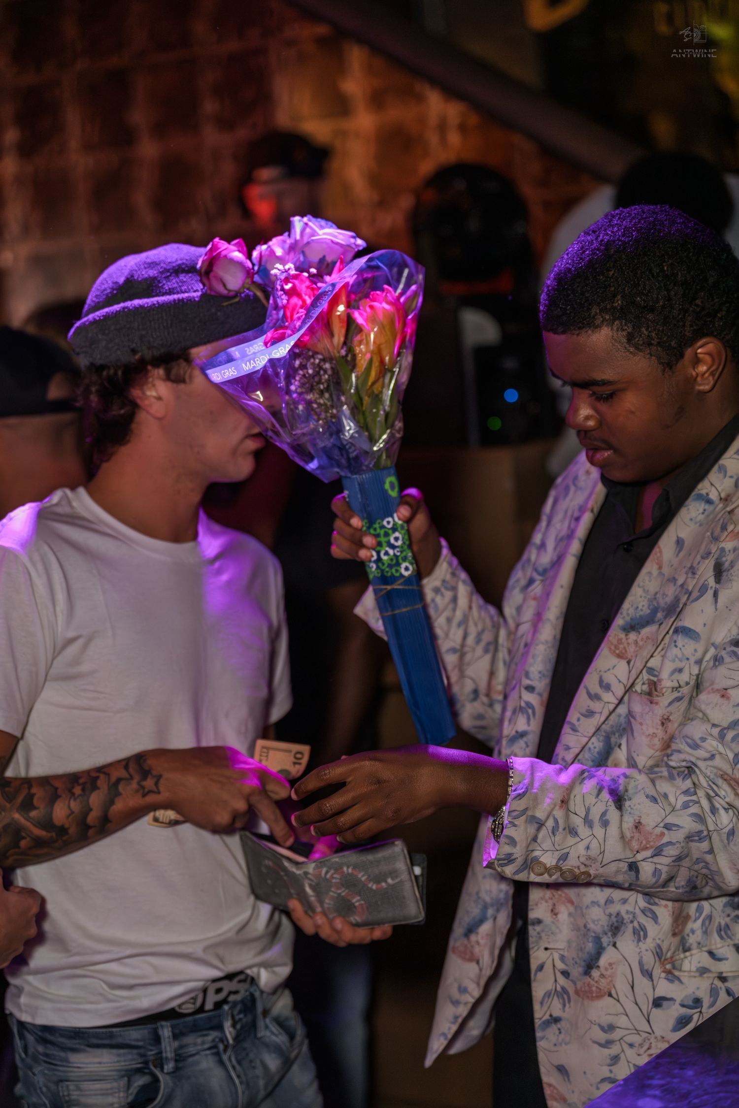 Two men trading a colorful flower bouquet under purple lights at a nightclub party.