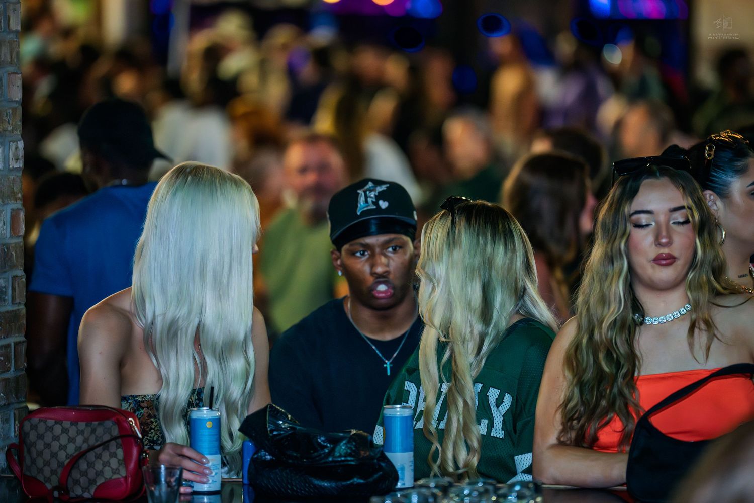 Man in black cap socializing with women at a busy nightclub bar.