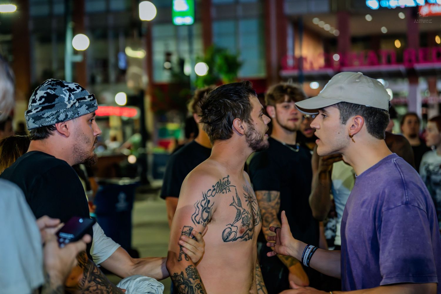 Shirtless tattooed man conversing with friends on busy city street at night.