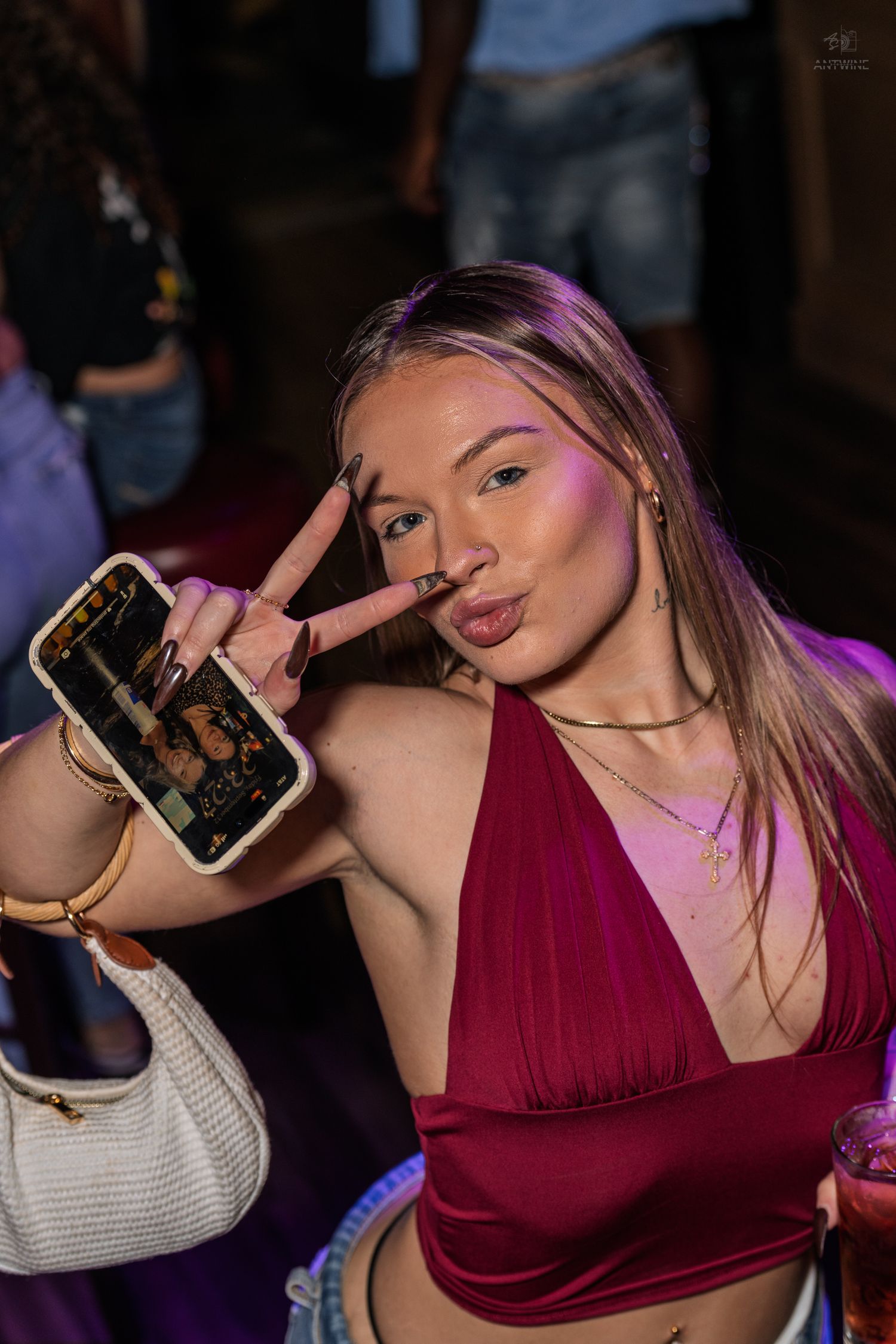 Young woman in red dress makes peace sign and pouty face while holding phone at a nightclub event.