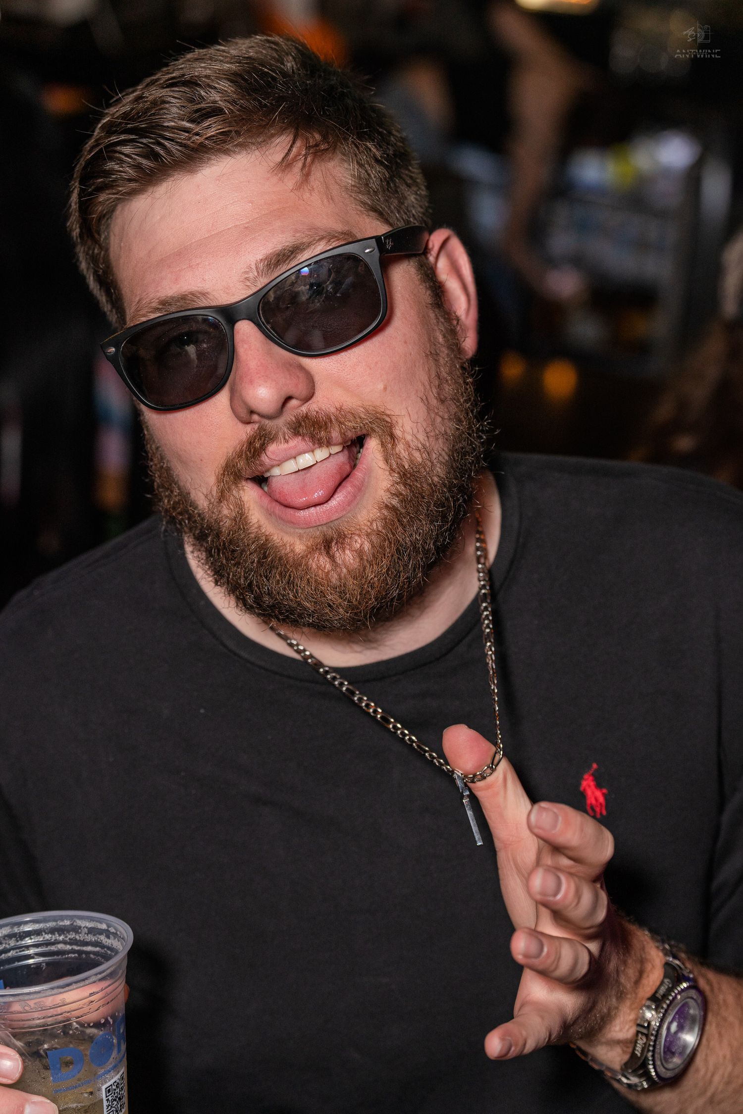 Bearded man in black shirt and sunglasses smiling and holding a drink at a nightclub.