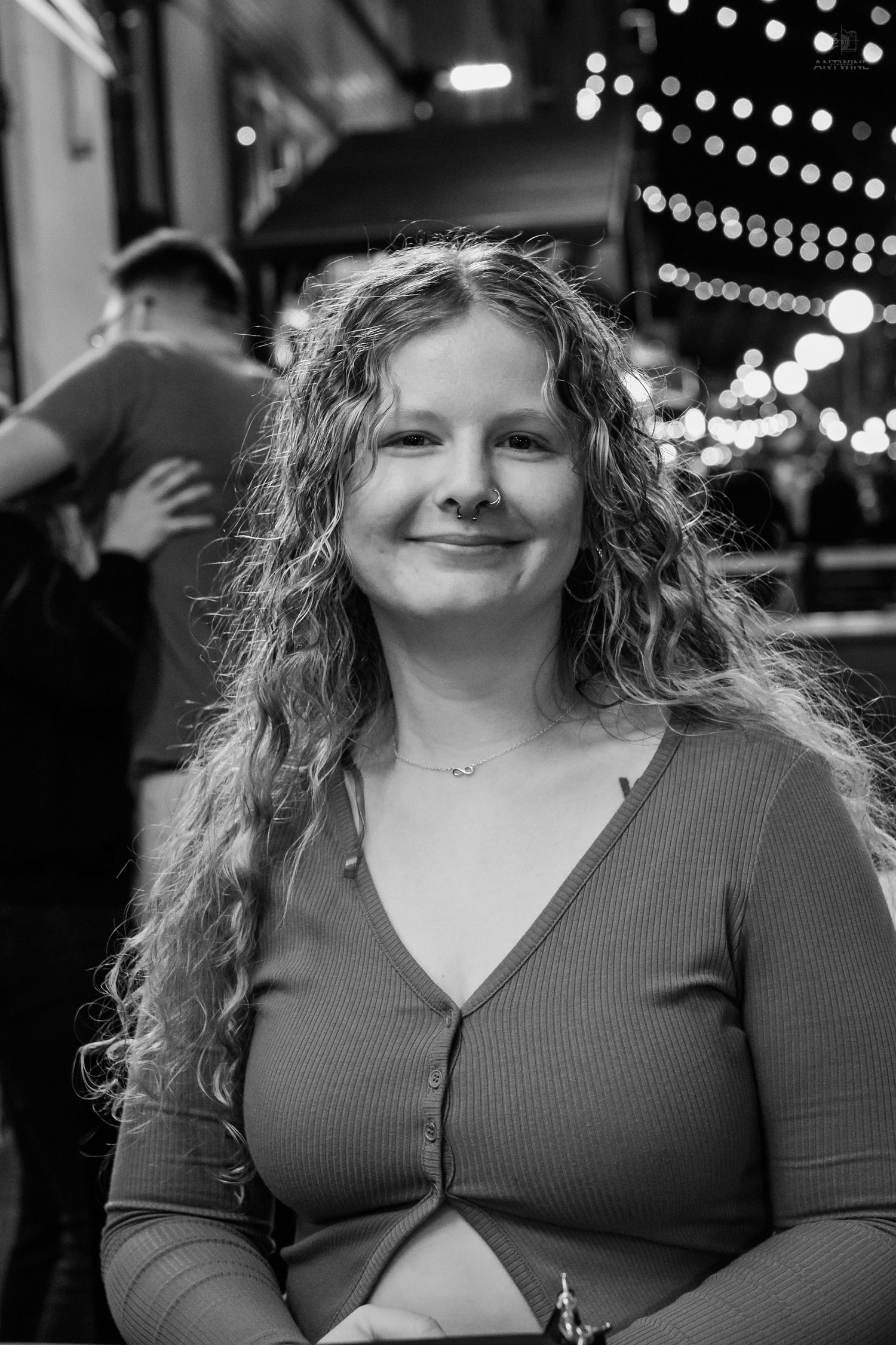Black and white portrait of a smiling curly-haired woman outdoors at night with bokeh lights behind.