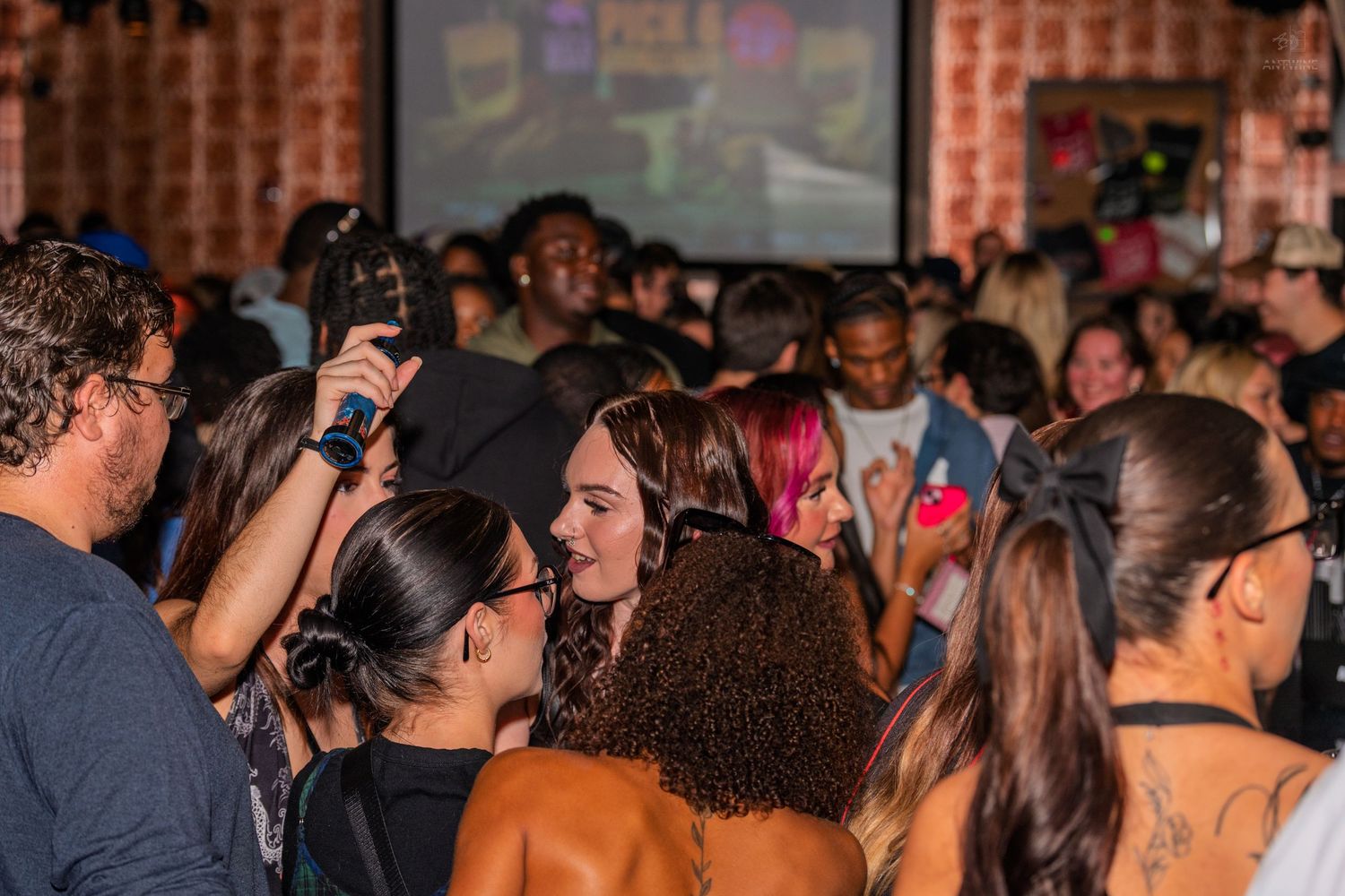 A diverse crowd mingles and smiles inside a packed nightclub with a projection screen.