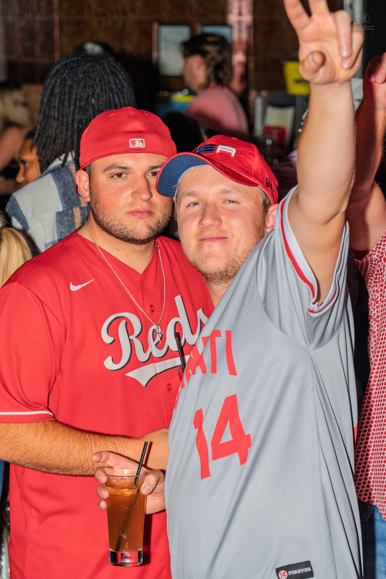 Two men in Cincinnati Reds jerseys posing at a crowded bar event.