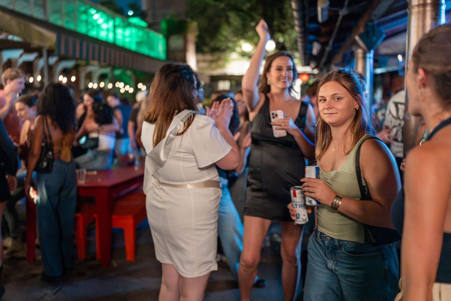 Women socializing at an outdoor nightlife event, holding drinks and dancing under string lights.