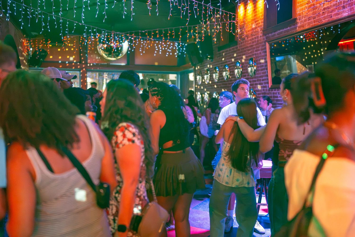 Crowded nightclub dance floor with colorful string lights and neon glow, people dancing and socializing at a lively party.