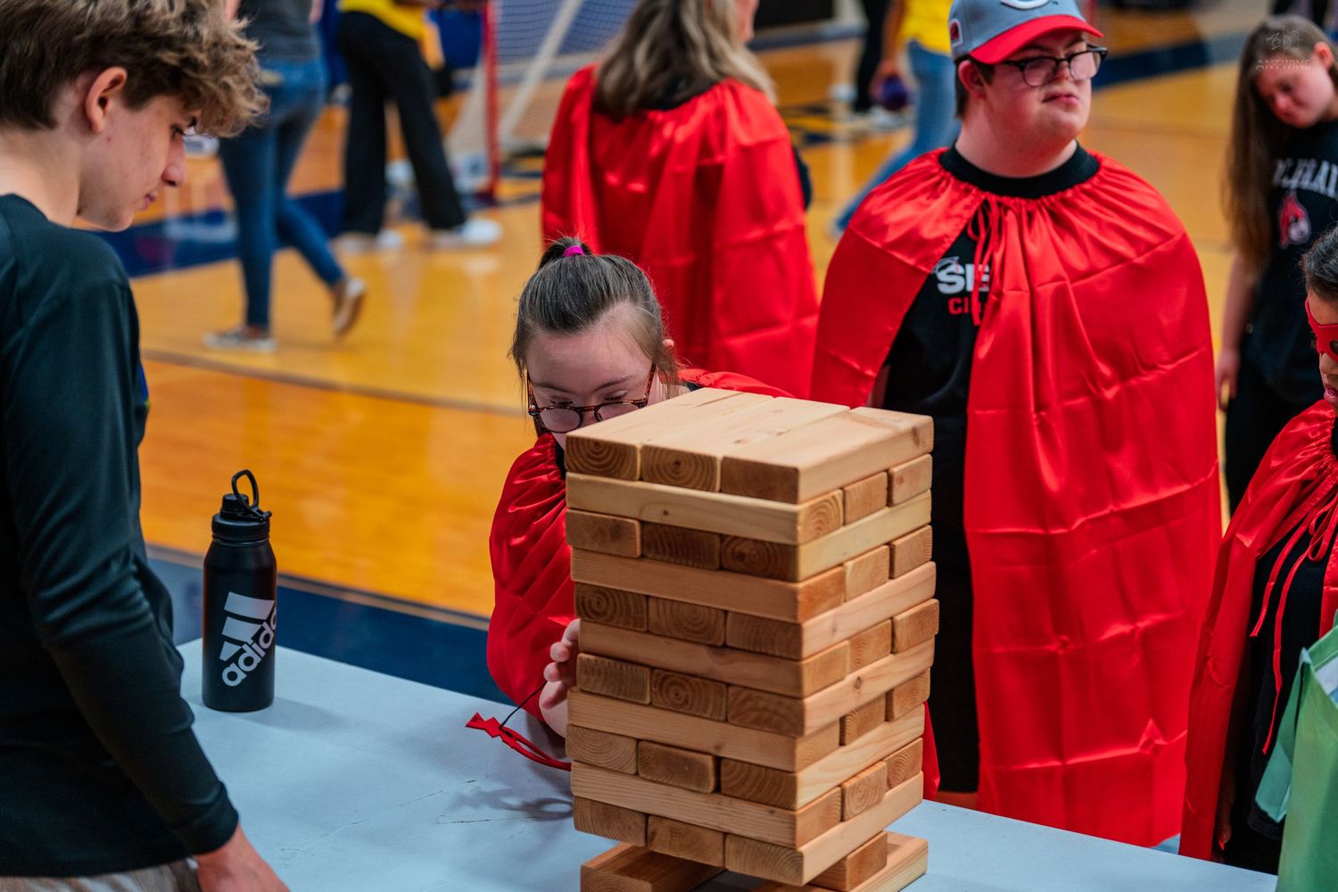Children in red capes play a giant Jenga game at an indoor school event on a gymnasium floor.