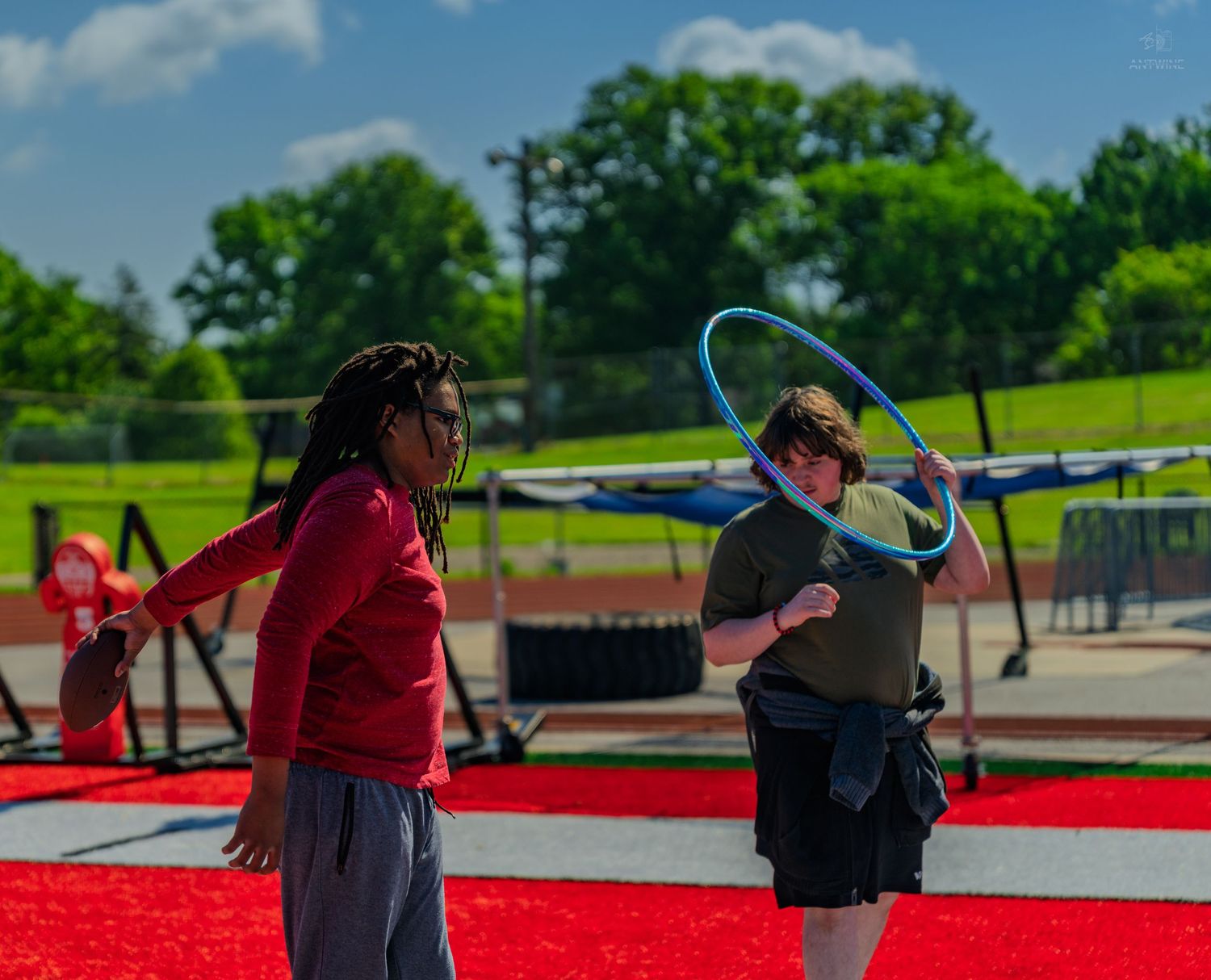 Two people playing with a hula hoop outdoors on a red track field on a sunny day.
