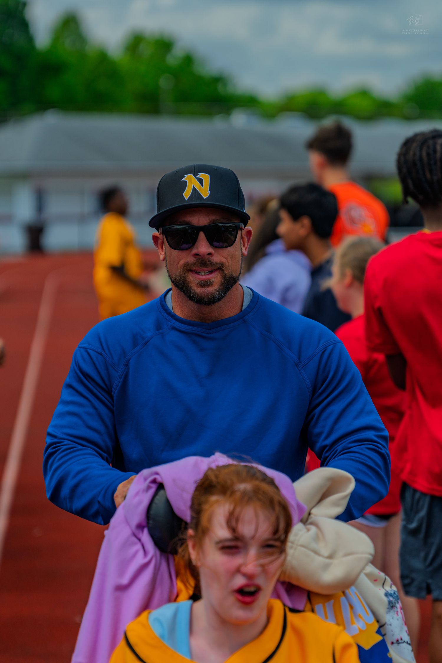 Man in blue shirt and N cap with sunglasses stands behind girl at outdoor track event.