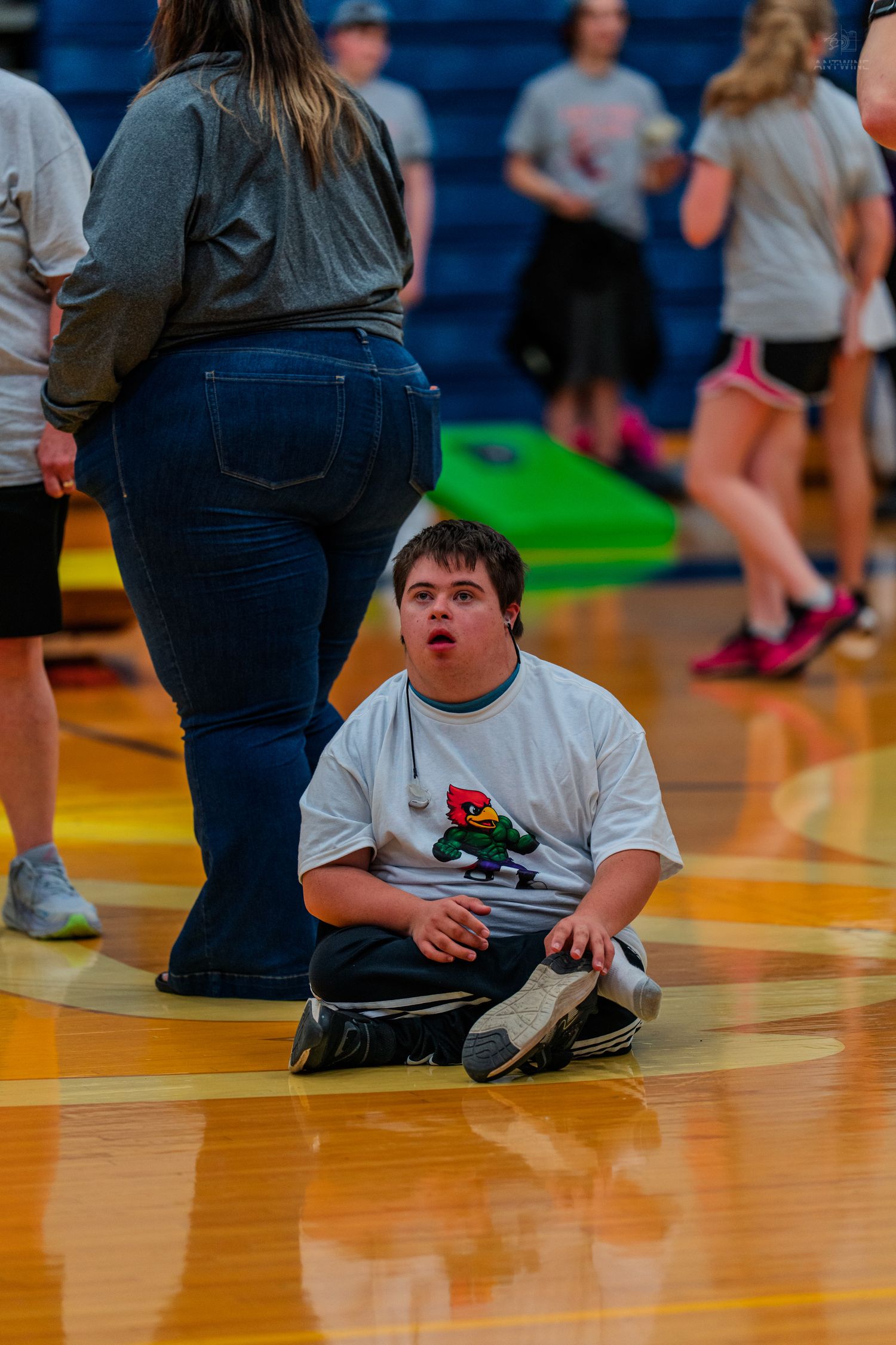 Child with Down syndrome sits on gymnasium floor during an inclusive indoor sports activity event.