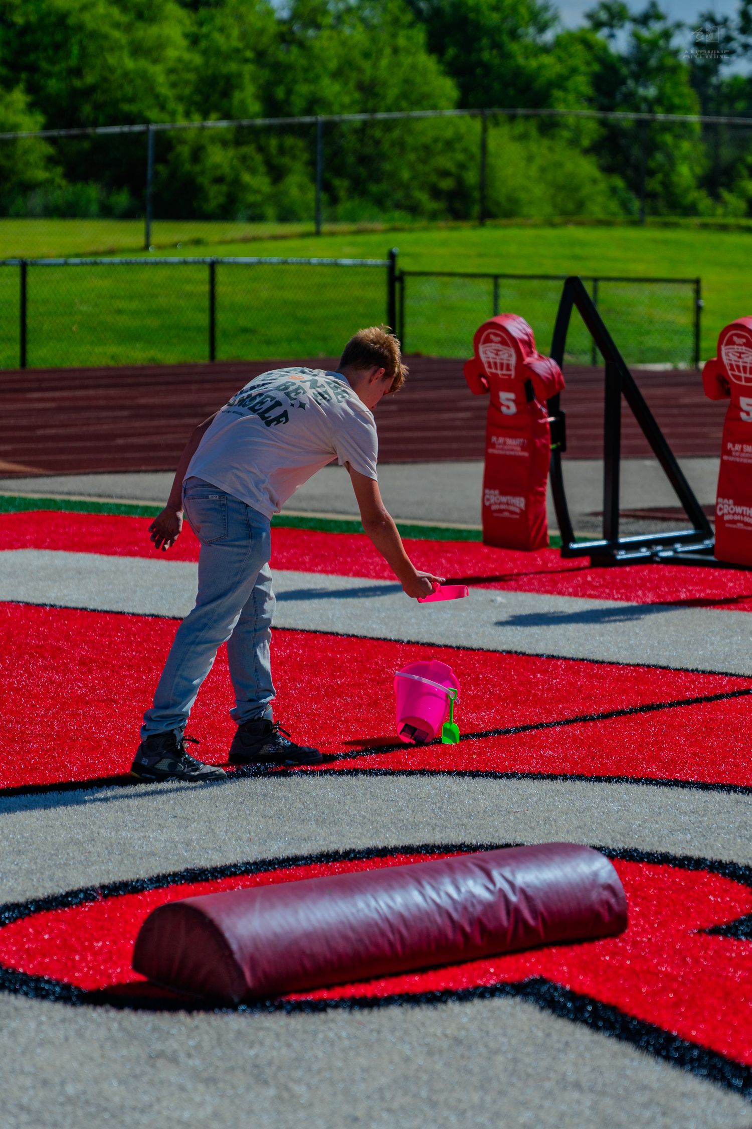 Person maintaining red artificial turf field with foam roller and pink tools near stadium track.