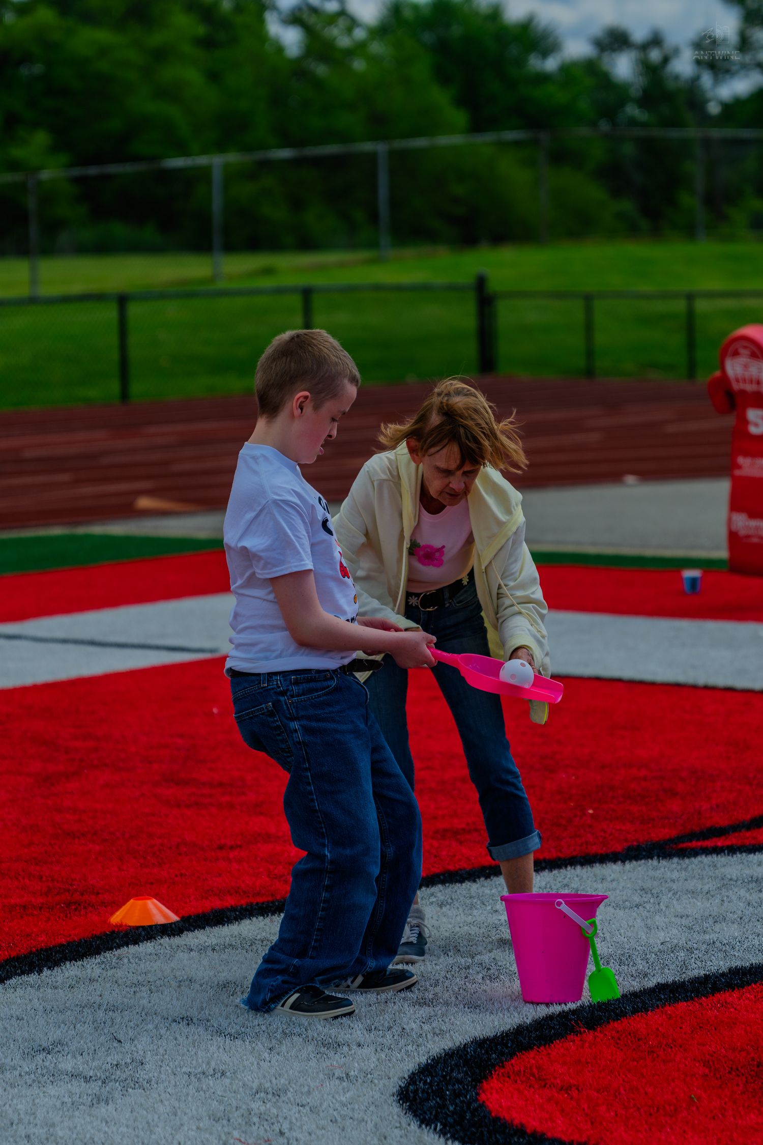 Two children playing with pink shovel and bucket on red turf field near running track outdoors.
