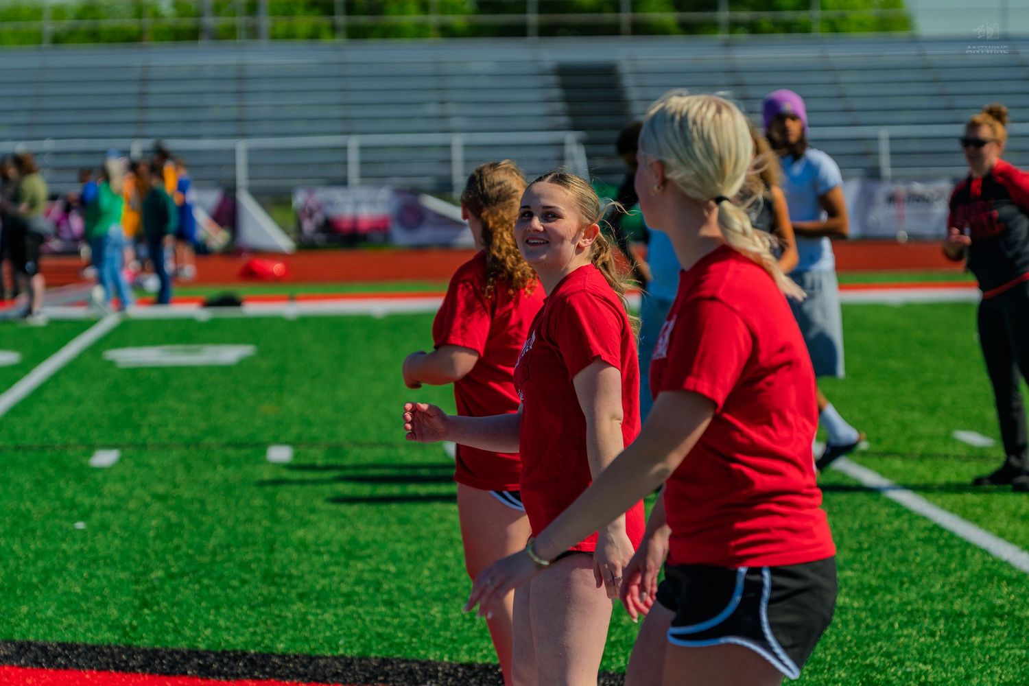 Young women in red shirts playing on a green turf athletic field during an outdoor sports event.