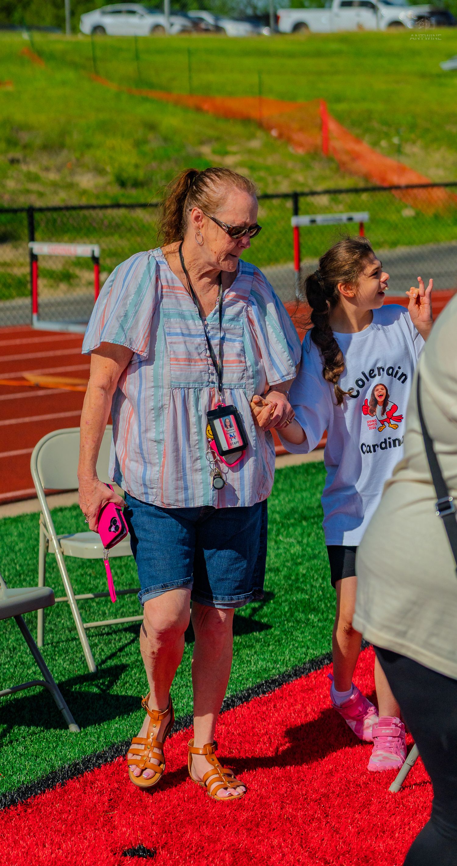 Woman with lanyard and girl in Cardinals shirt stand on red turf near a track field outdoors.