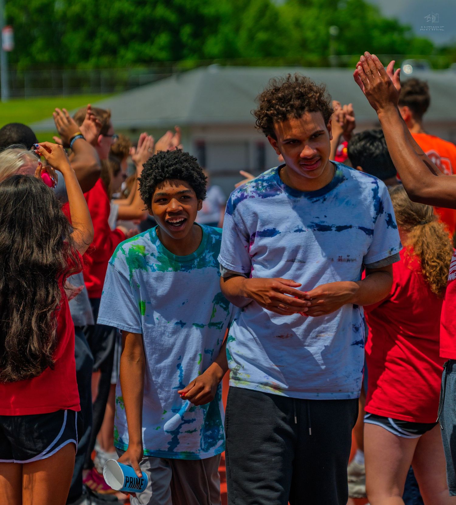Two smiling teens in tie-dye shirts celebrate outdoors at a colorful school event with peers in red shirts.