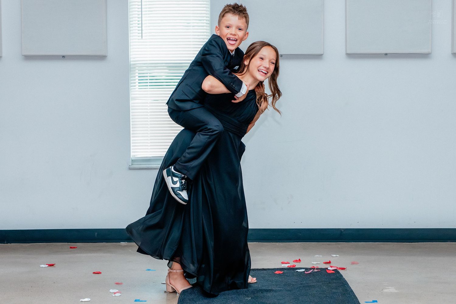 Woman in black gown giving a young boy a piggyback ride at a wedding celebration indoors.