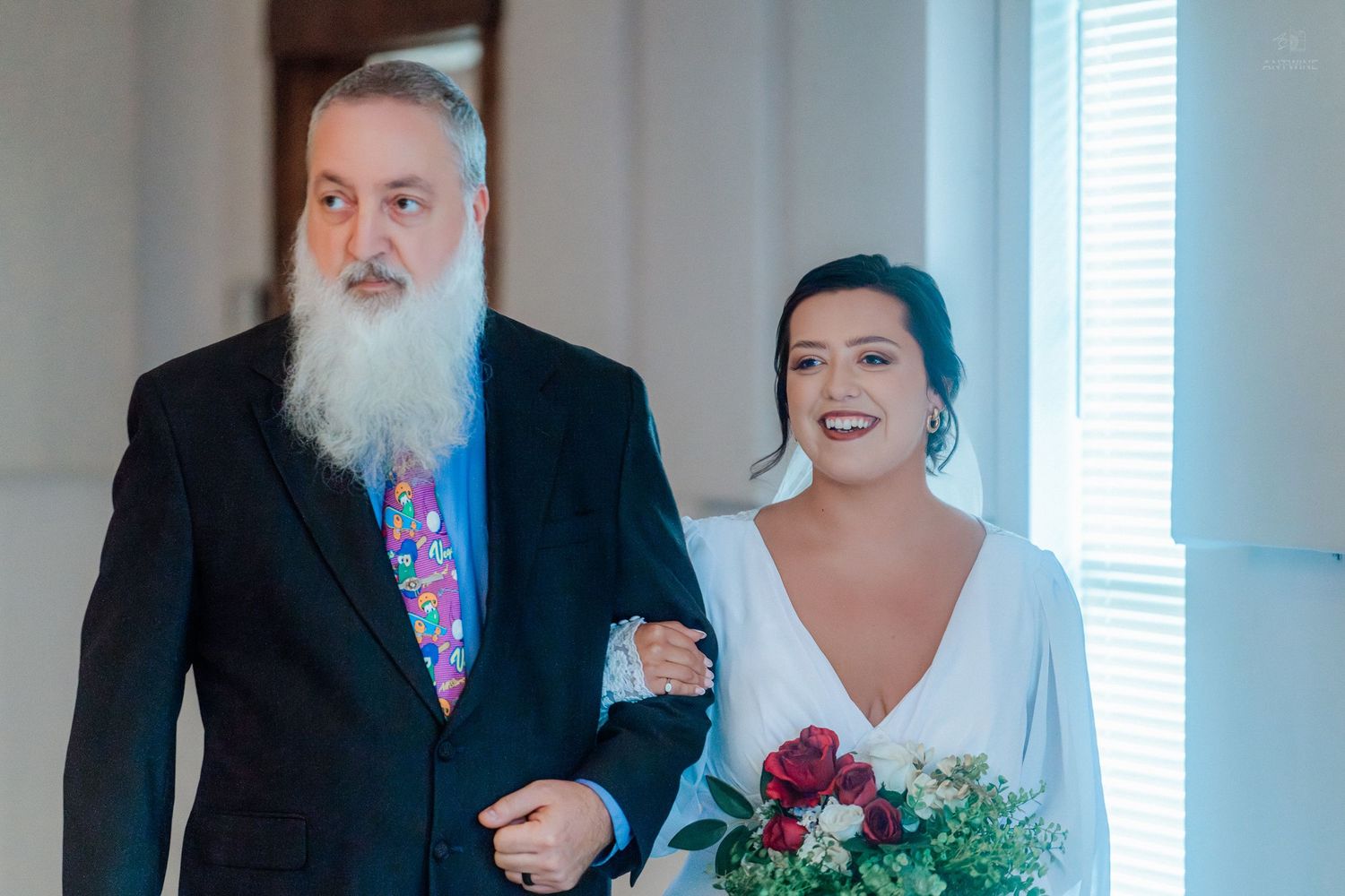 Bride in white gown walks down the aisle with bearded man in dark suit and floral tie holding red bouquet.