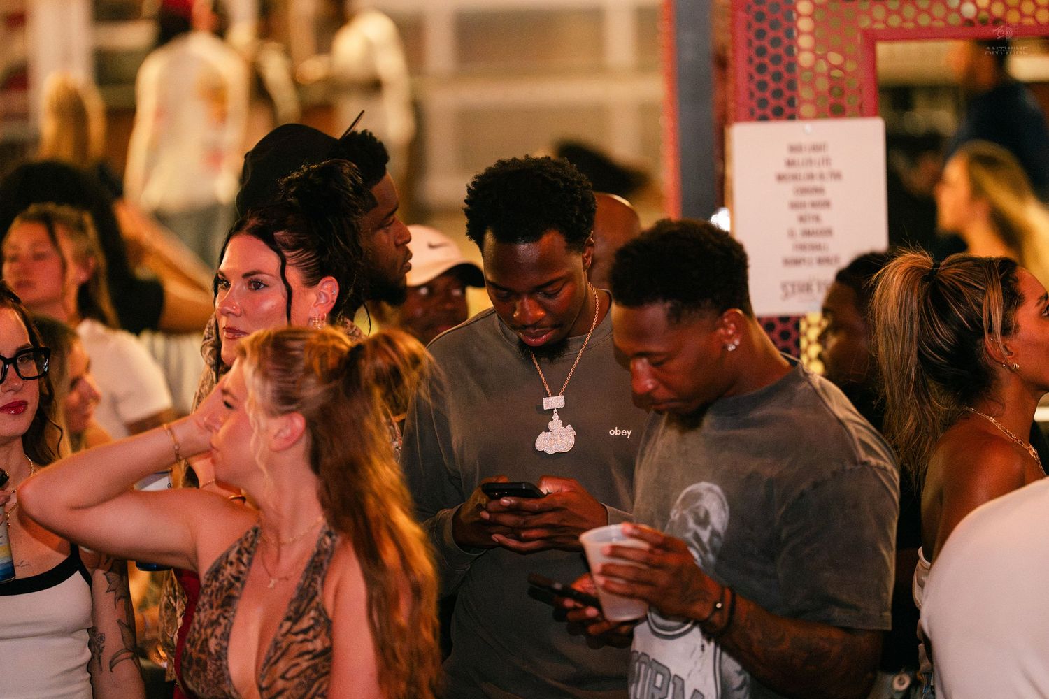 Young men checking phones in a crowded outdoor nightlife venue surrounded by a diverse group of partygoers.