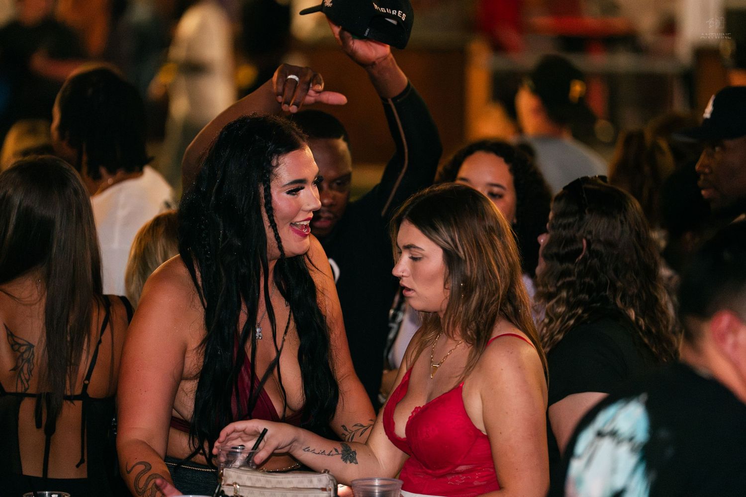 Two women in elegant outfits chat at a lively nightclub event.
