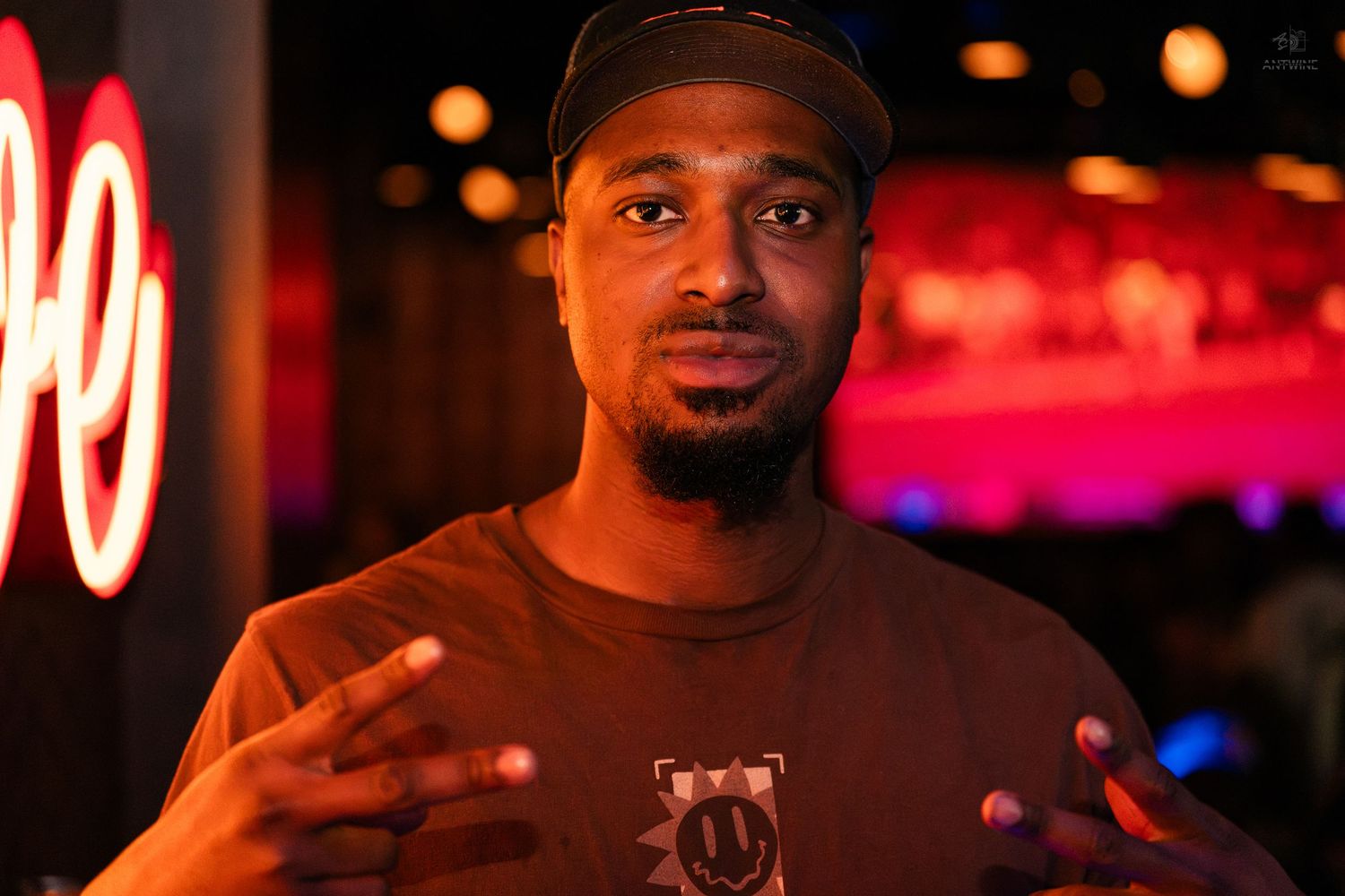 A man in a brown shirt and cap gestures with both hands at a neon-lit venue.